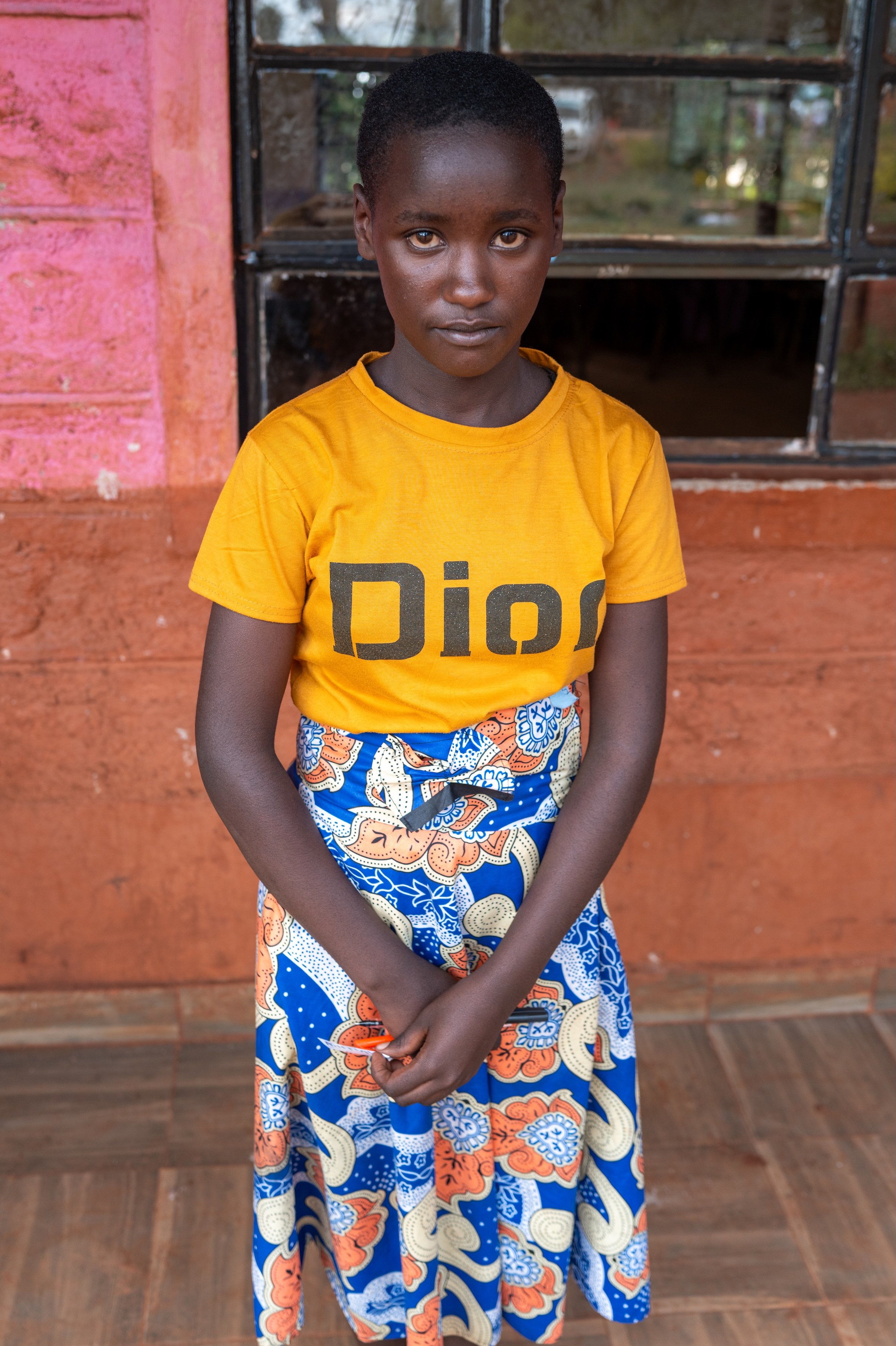 A young girl with dark skin and short hair stands indoors in front of a window and a colorful wall. She is wearing a yellow T-shirt and a long, patterned skirt. Her hands are clasped in front of her and she looks directly at the camera.