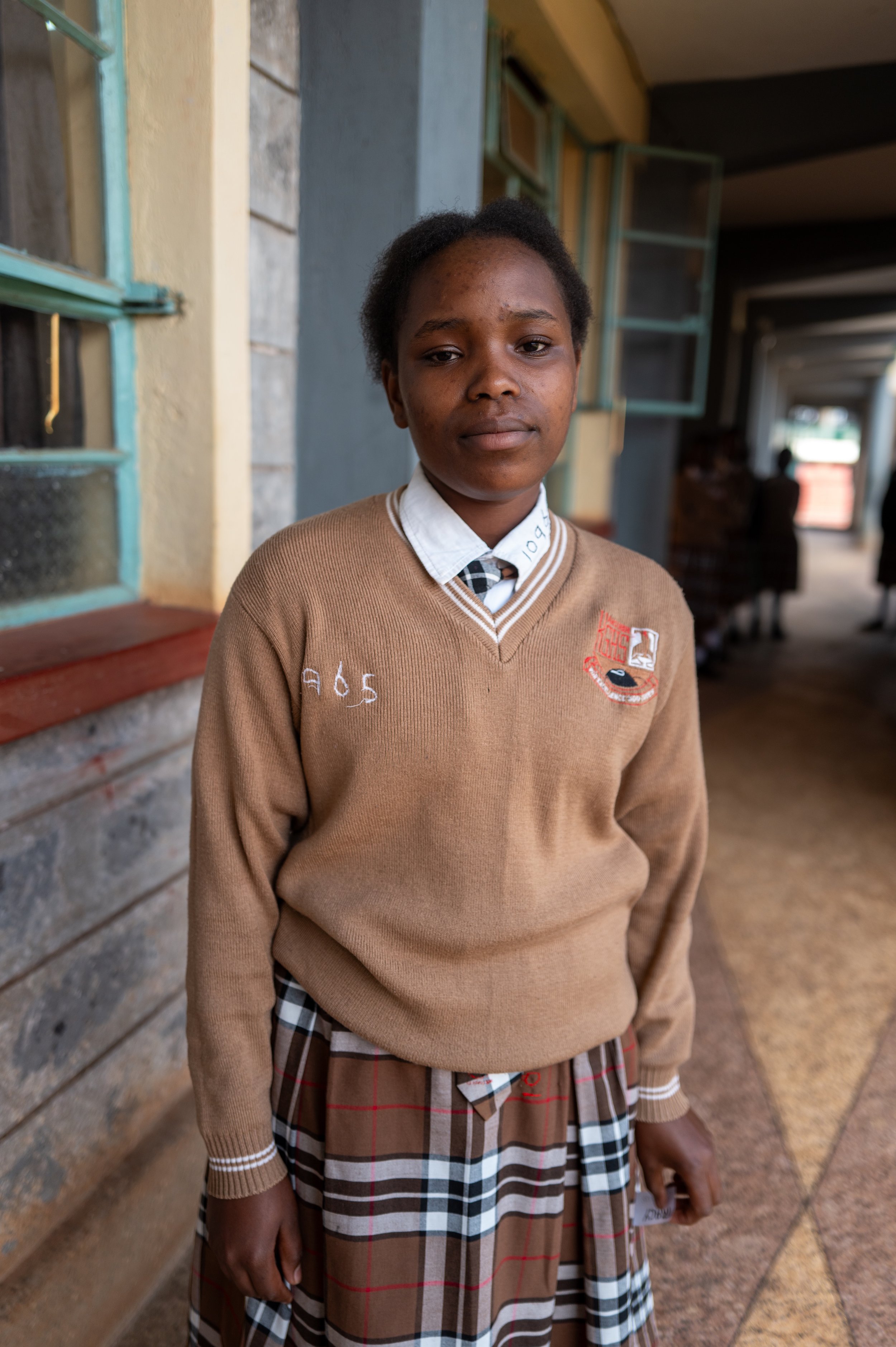 A young girl in a school uniform standing in a corridor with windows and other students in the background.