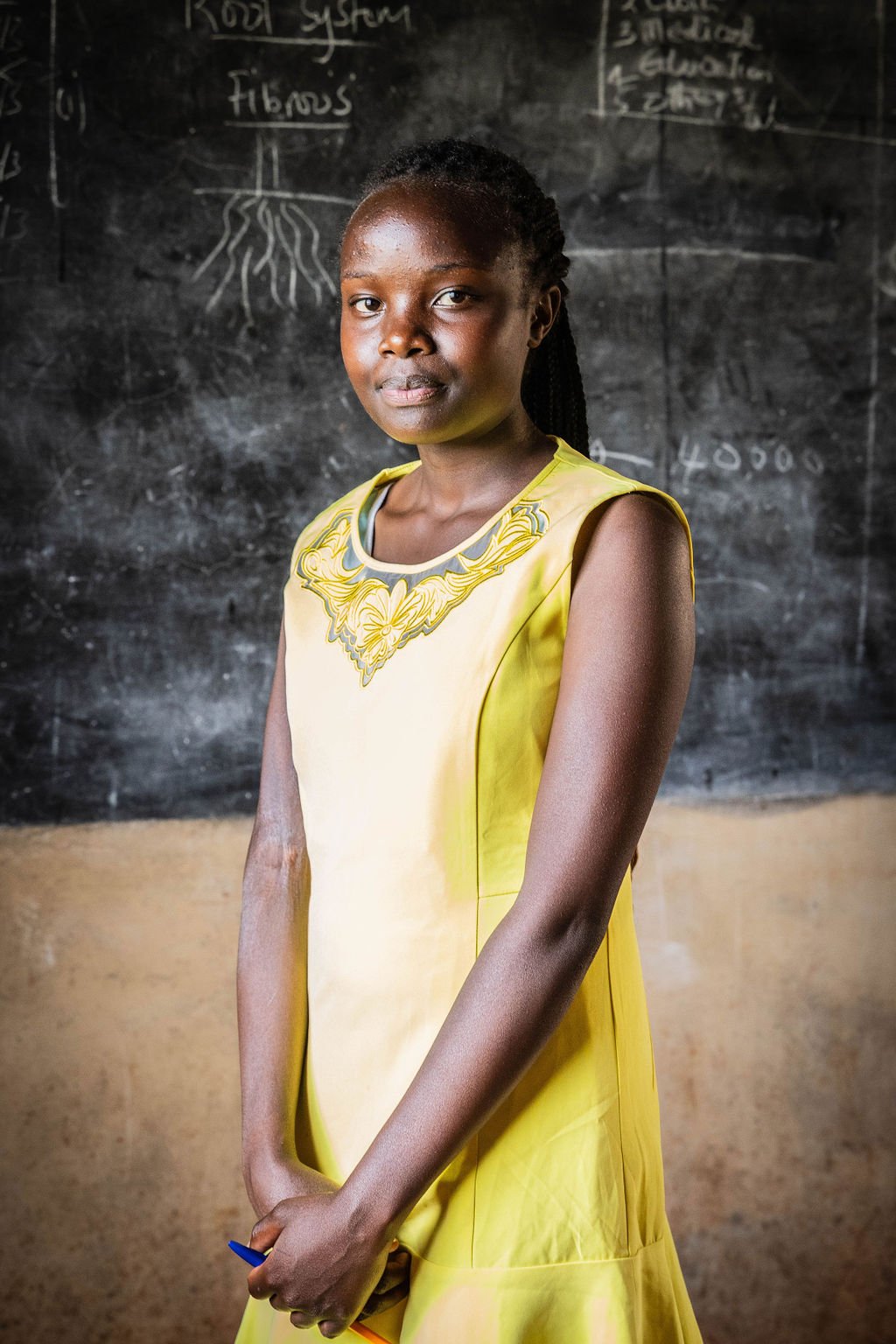Young girl in a yellow dress stands in front of a blackboard with diagrams.