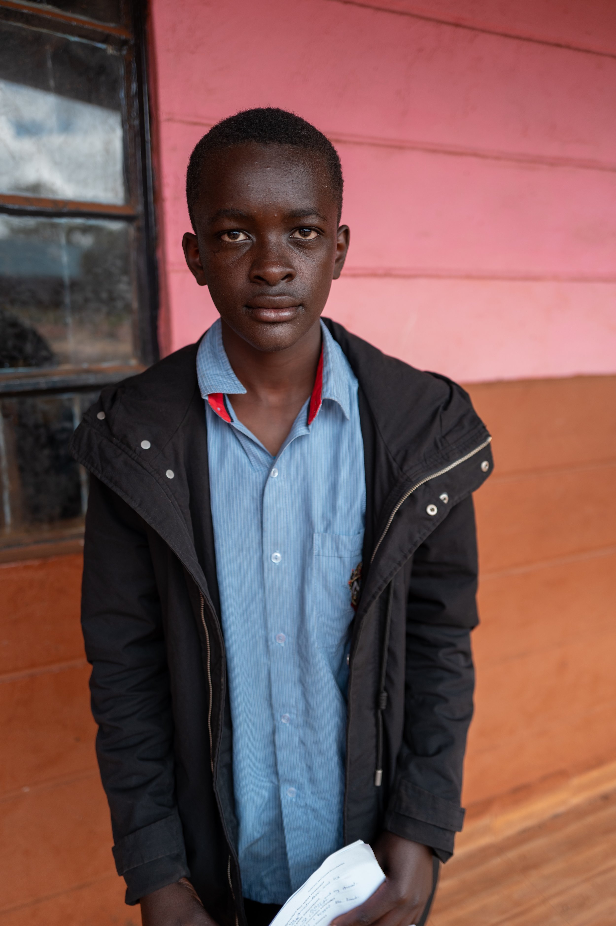 A young boy wearing a blue shirt and black jacket, standing indoors in front of a pink wooden wall, holding a piece of paper.