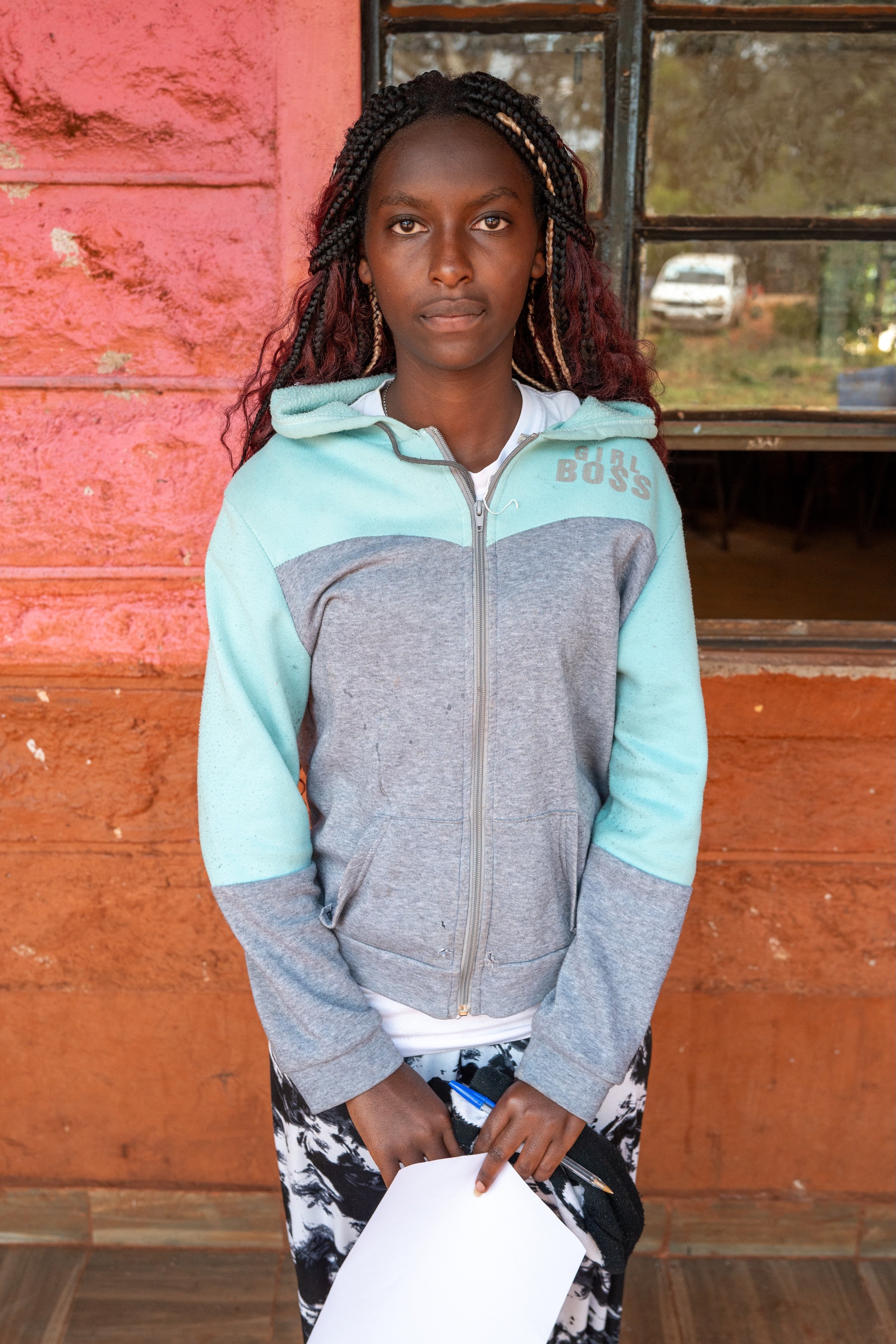 A young girl with braided hair standing in front of a rustic red wall with a window behind her. She wears a light blue and gray zip-up hoodie, patterned pants, and holds a piece of paper with a pen in her hand.