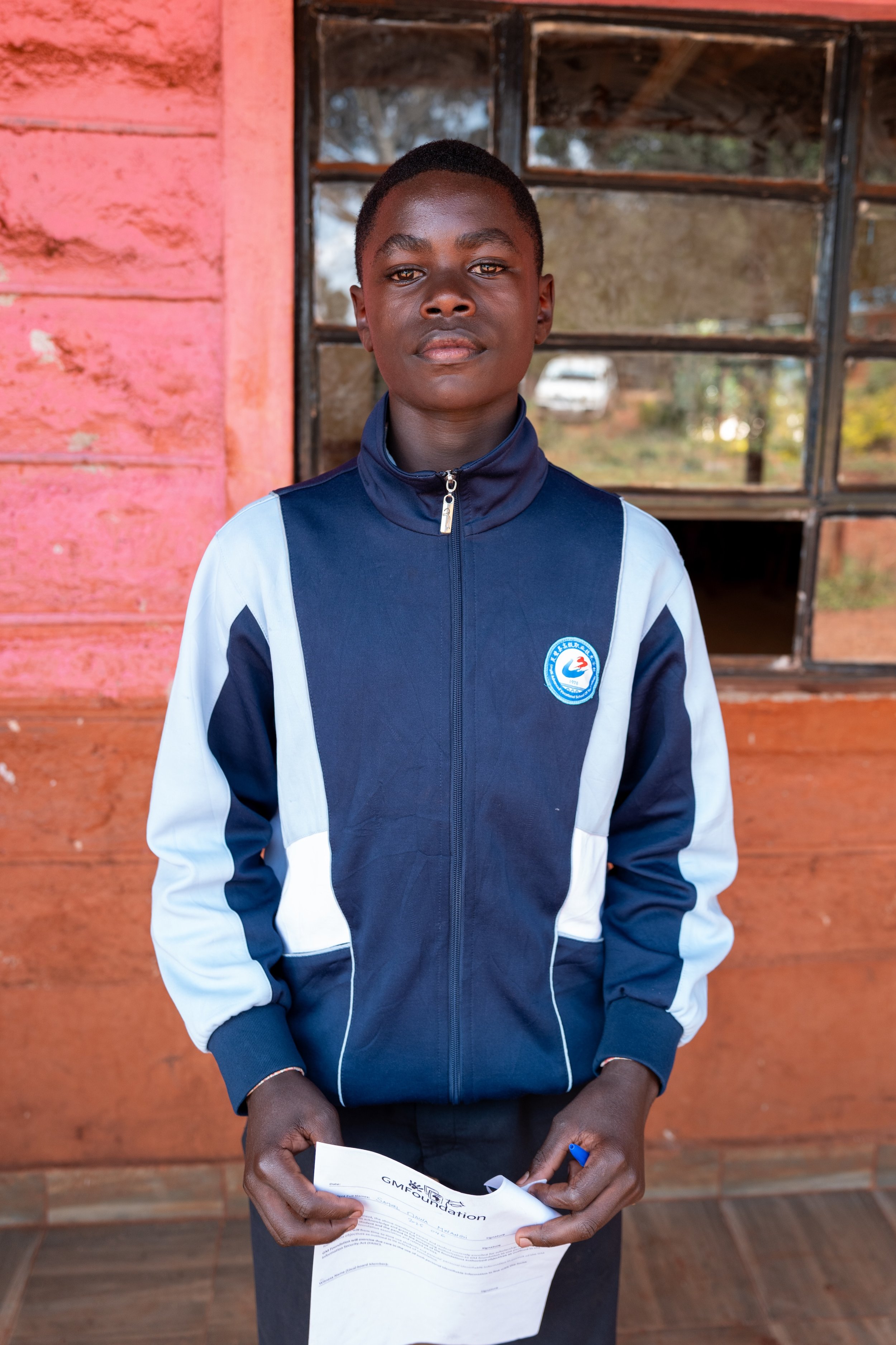 A young boy standing in front of a wooden wall with a window, holding a piece of paper, wearing a blue and white sports jacket with a logo on the chest.