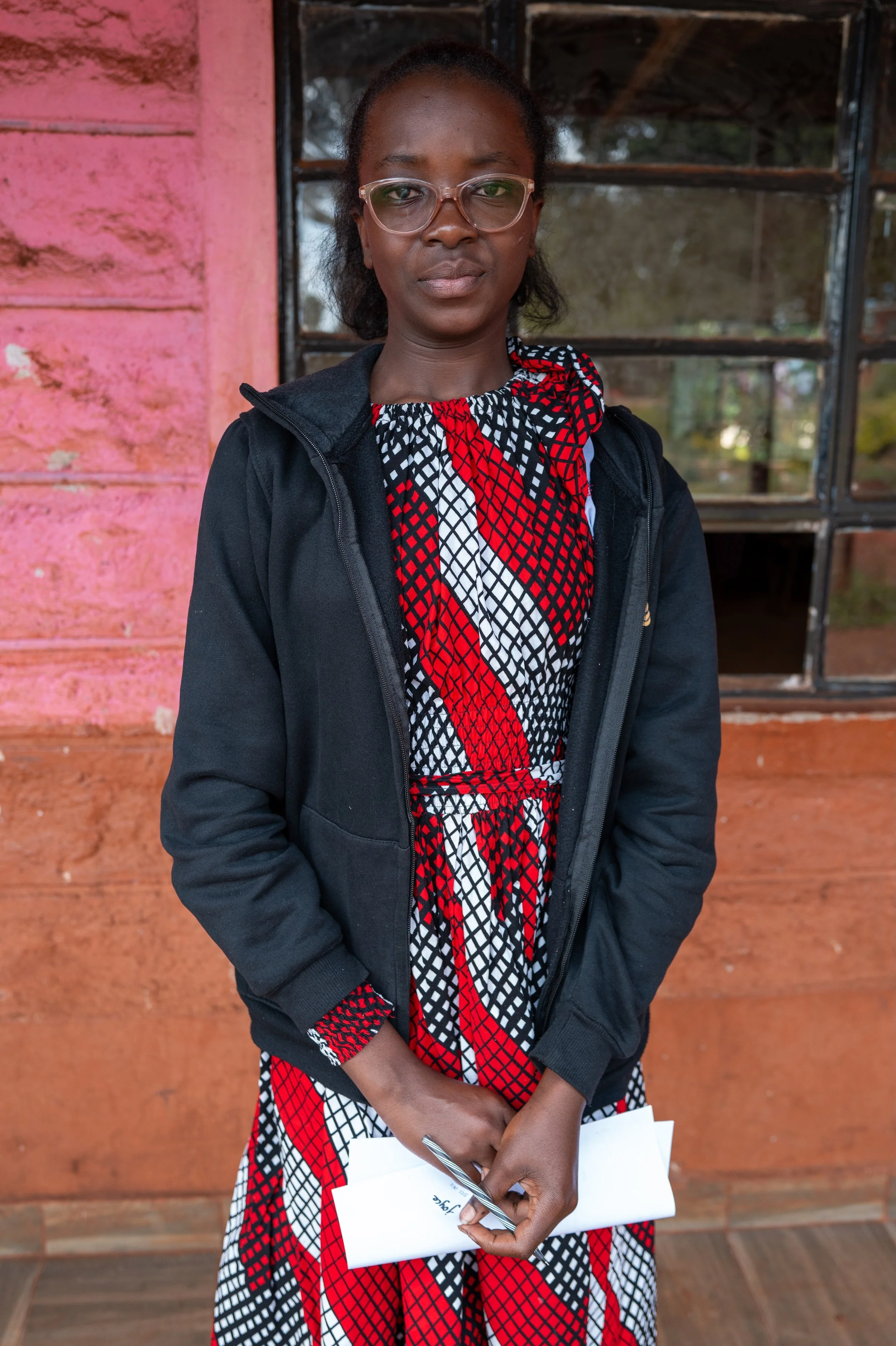 A woman wearing glasses, a black jacket, and a patterned dress holding a rolled-up piece of paper and a pen, standing outdoors in front of a pink wall and a window.