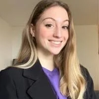 A young woman with long blonde hair smiling, wearing a black blazer and a purple top, against a neutral background.