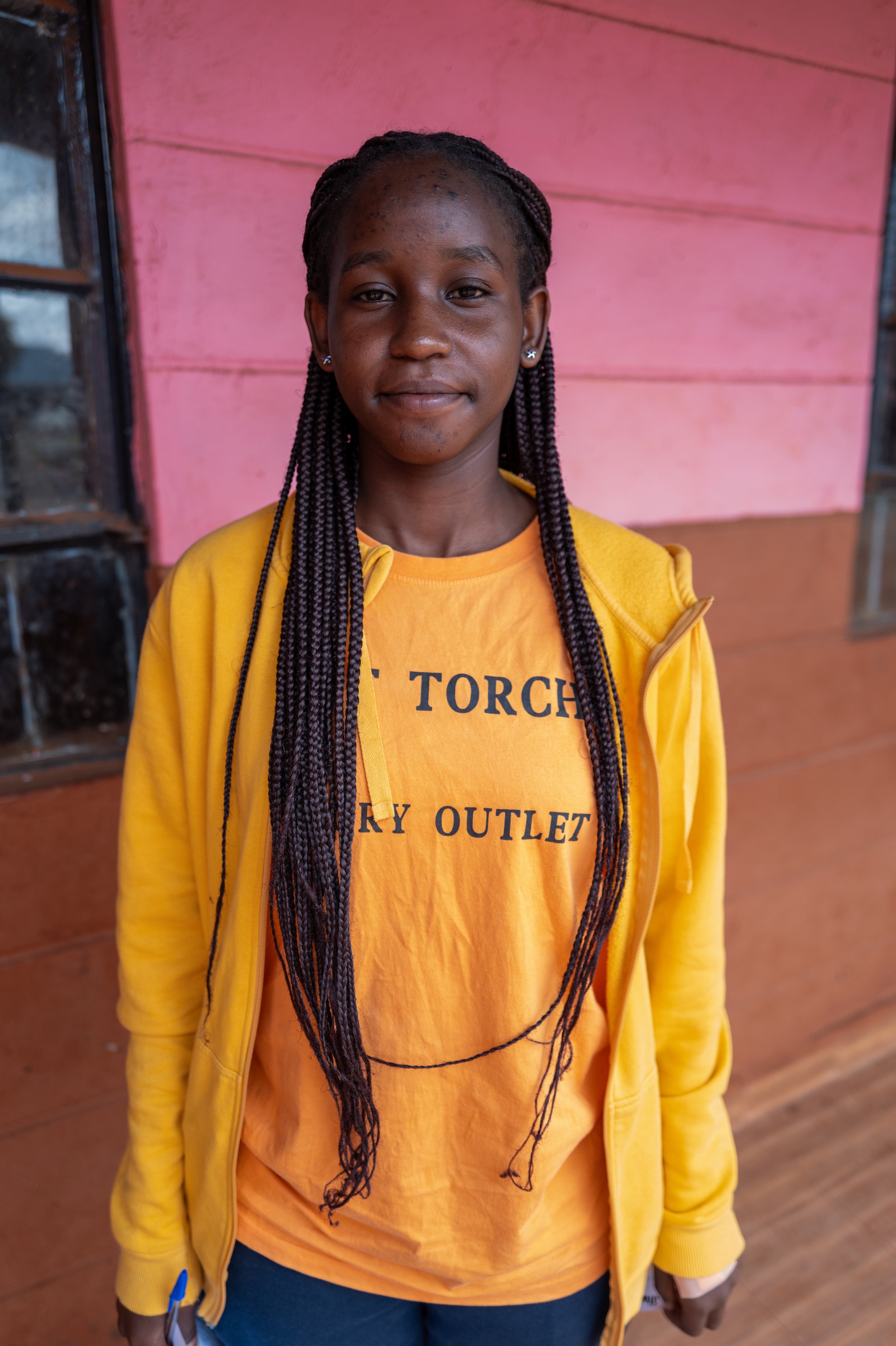 A young woman with braided hair, wearing a yellow jacket and an orange T-shirt that says 'TORCH SKY OUTLET,' standing in front of a pink wooden wall.