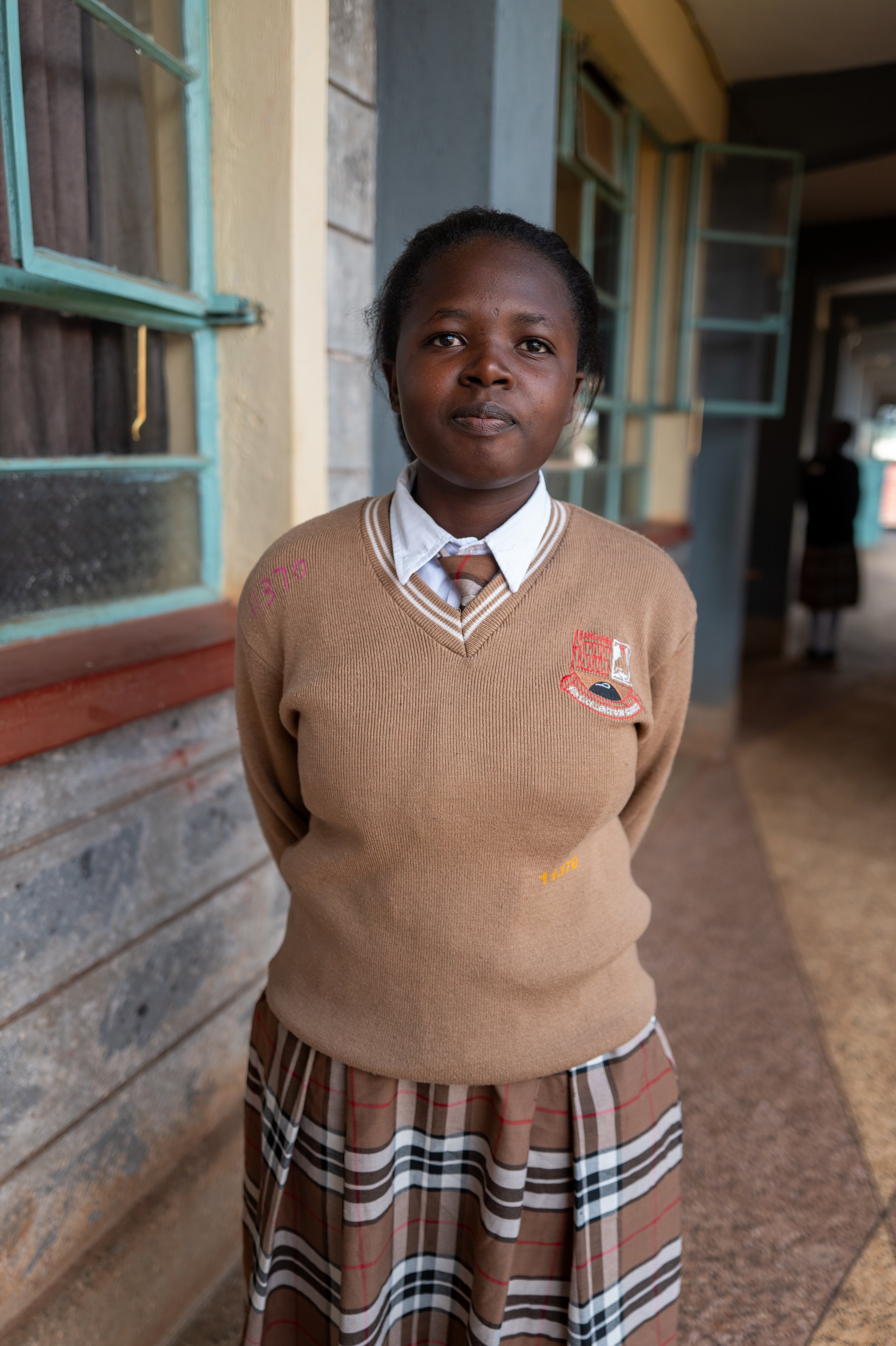 A young girl in school uniform standing in front of a brick and wood wall, with open windows, in a school corridor.