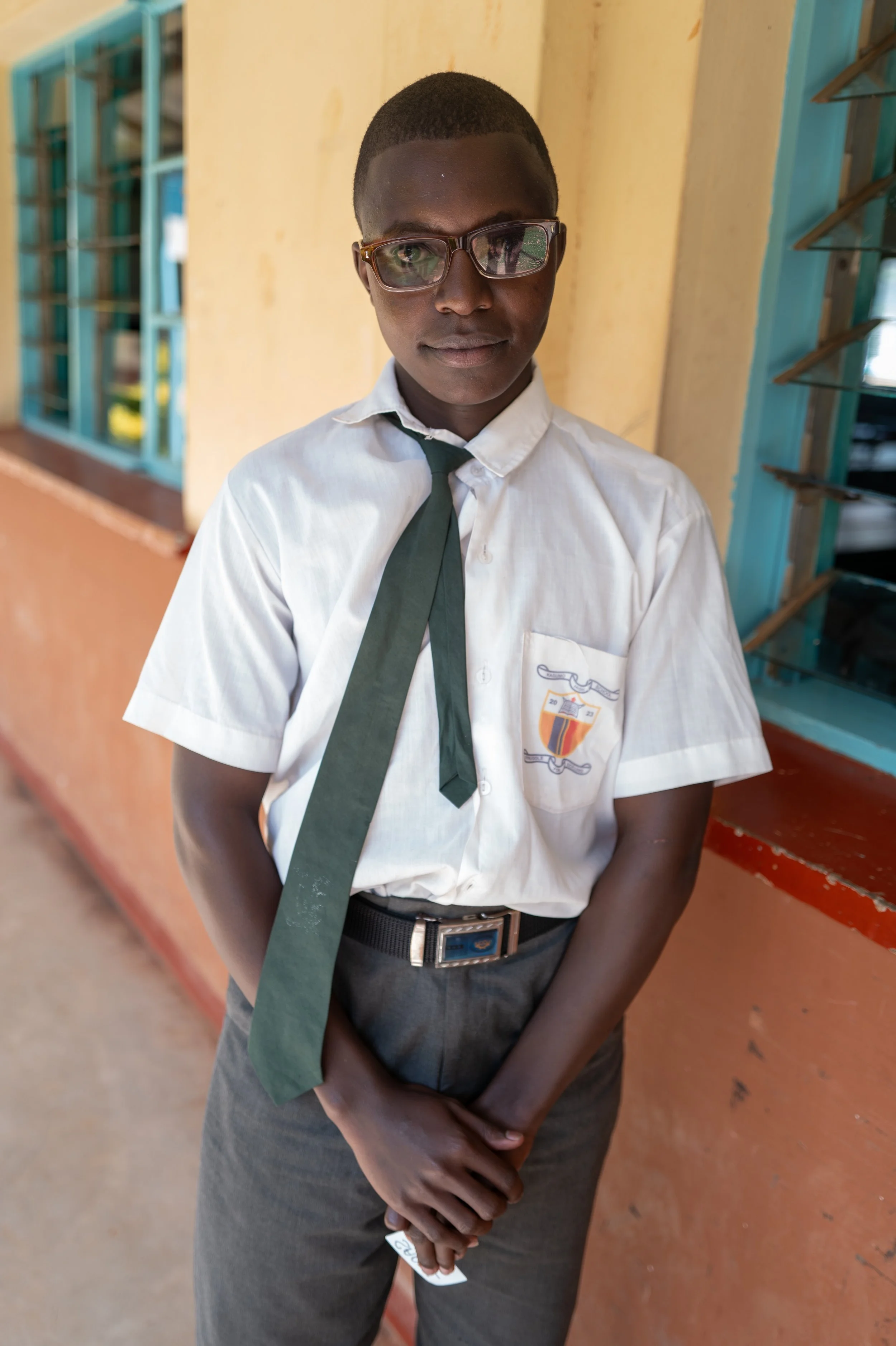 A young boy in a school uniform with glasses, standing indoors near a yellow wall with a window in the background.