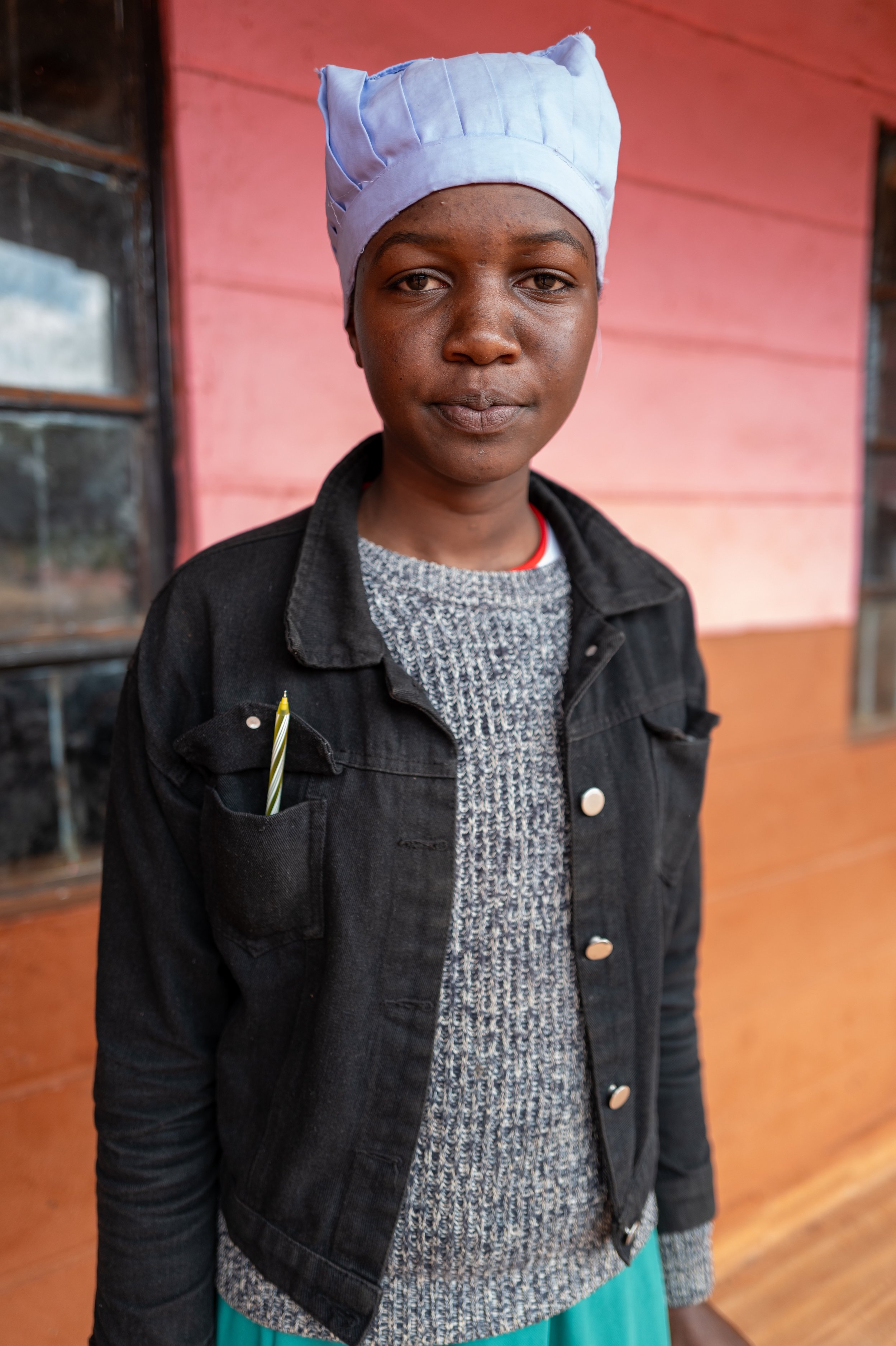 A young woman wearing a headscarf, a gray sweater, and a black jacket with a pen in her pocket, standing in front of a pink wooden wall.
