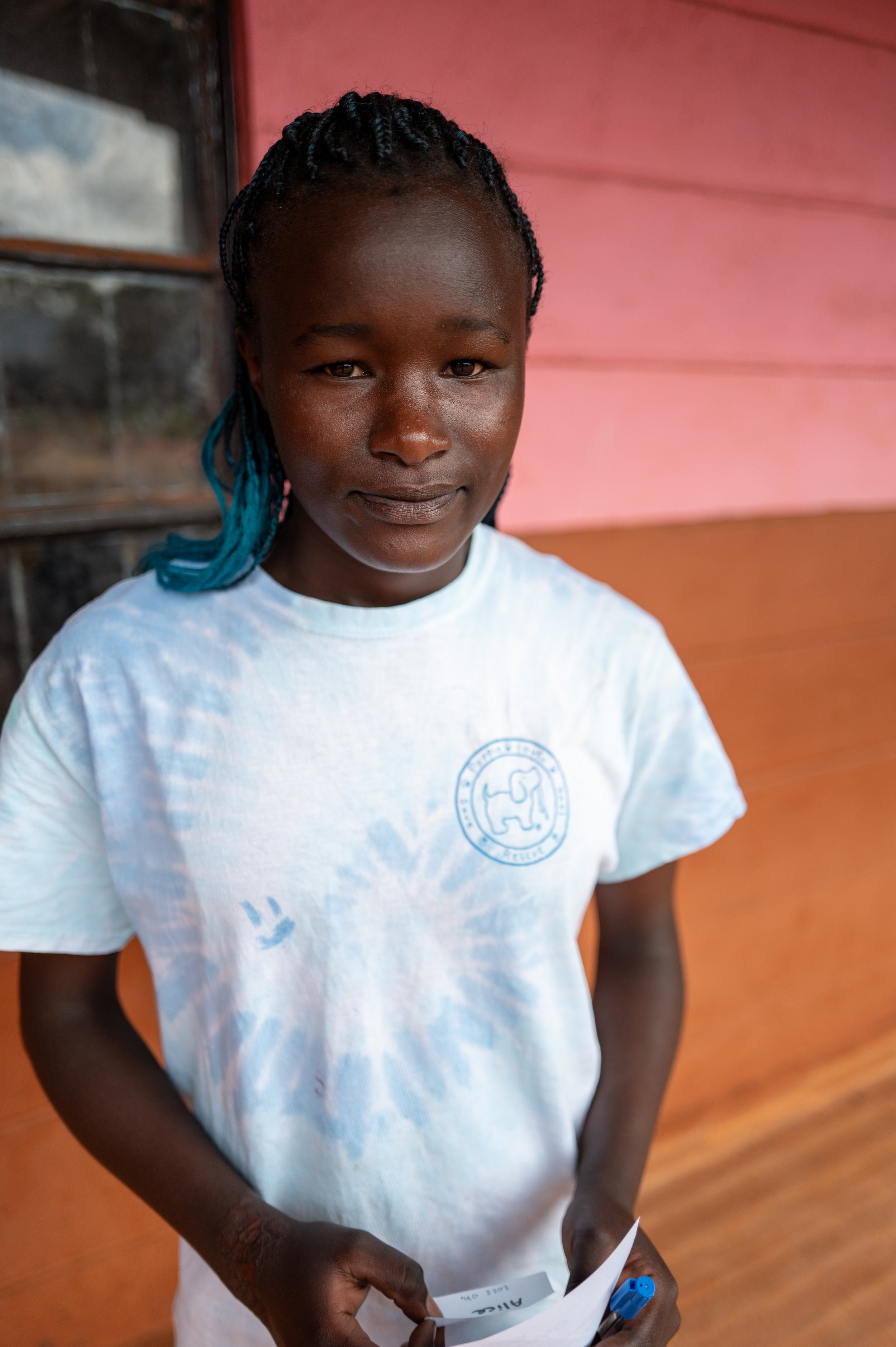 A young girl with braided hair and a slight smile, holding a piece of paper and a pen, standing against a pink and orange wall.