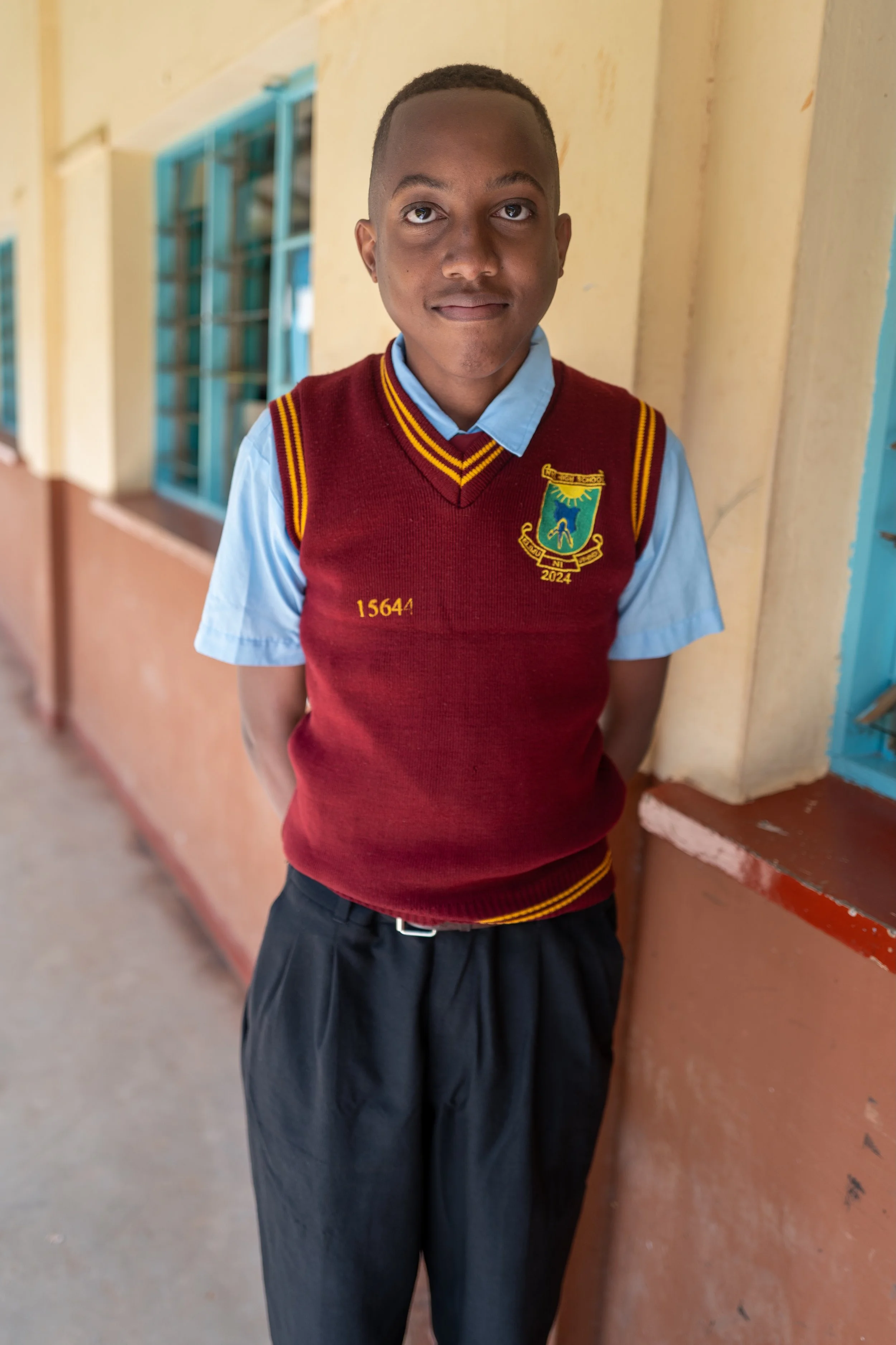 Young boy in school uniform standing by a building with yellow walls and blue window frames.