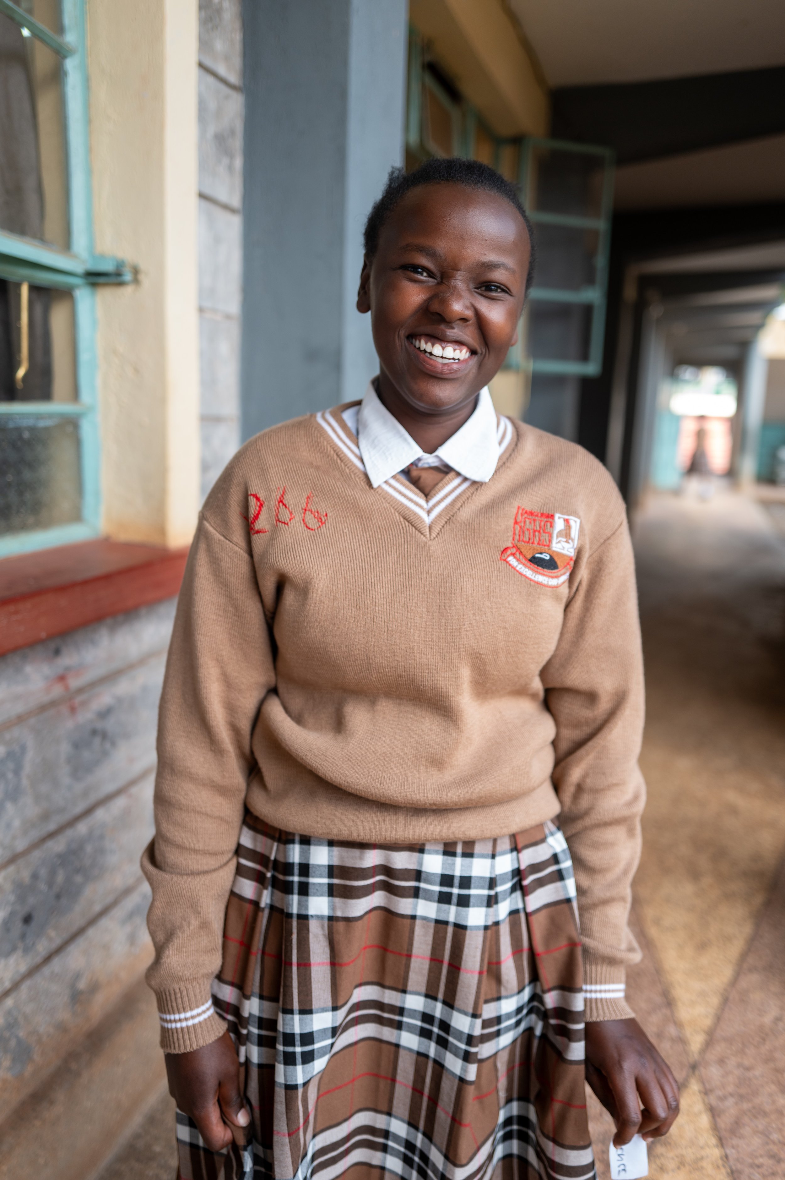 A smiling young girl in a school uniform standing outdoors next to a building with open windows.