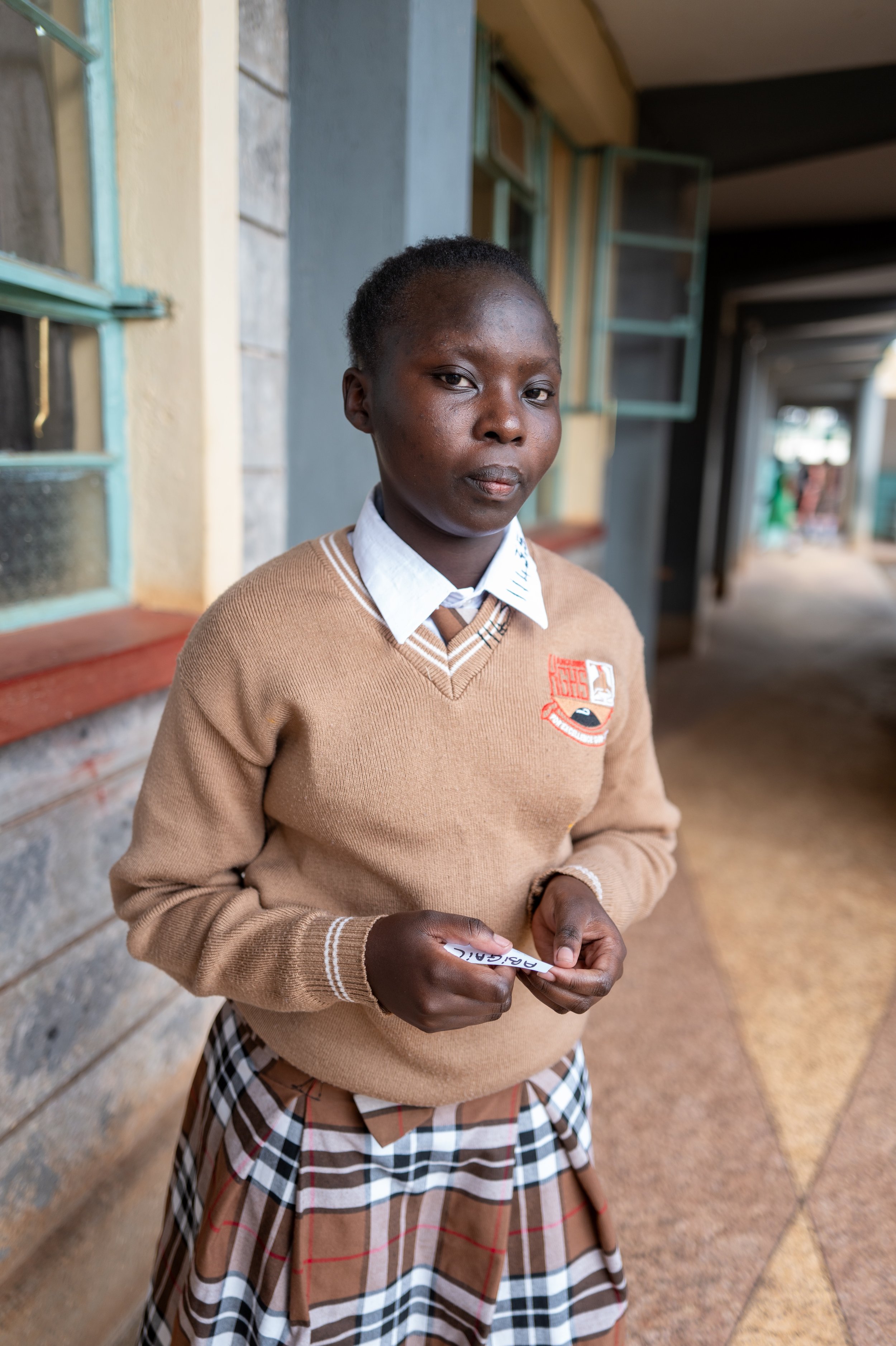 A young girl in a school uniform standing outside a school building, holding a name tag.