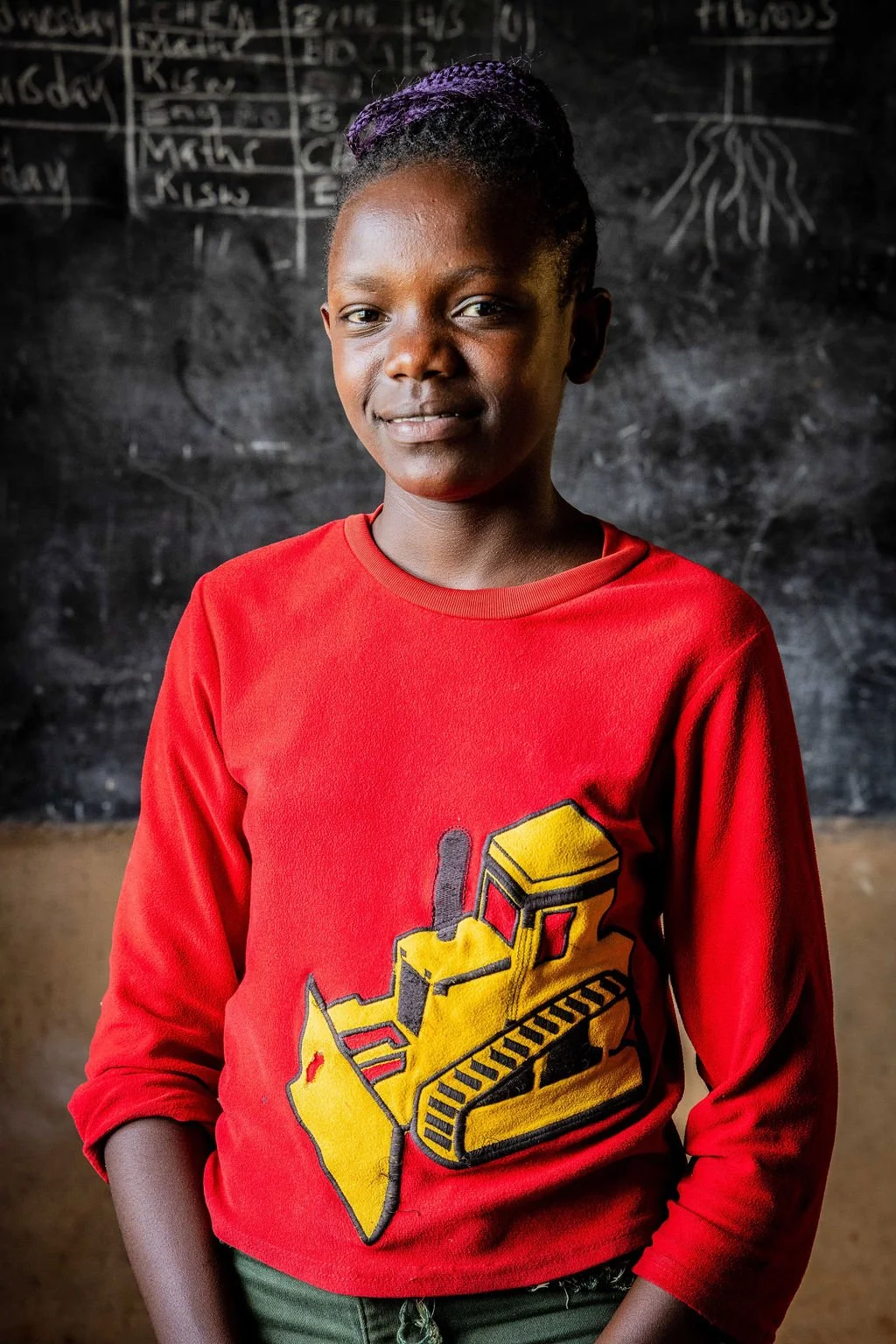Person wearing a red shirt with a yellow bulldozer graphic, standing in front of a chalkboard with writing.