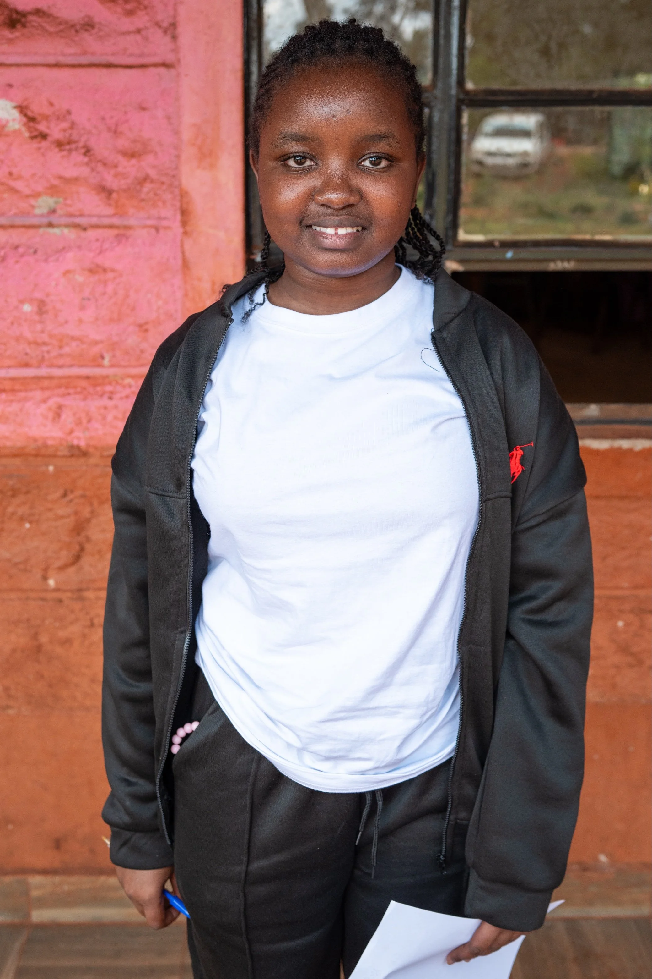 A young girl standing outdoors in front of a pink-painted wooden wall, wearing a white t-shirt, black jacket, and black pants, holding a piece of paper.