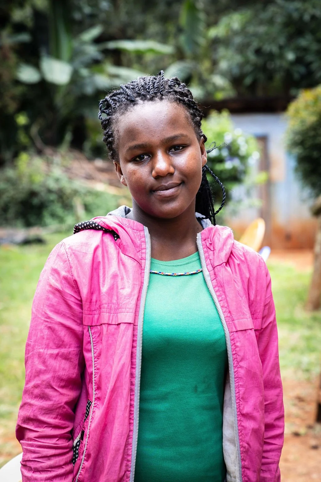 A person wearing a pink jacket and green shirt standing outdoors with a blurred leafy background.