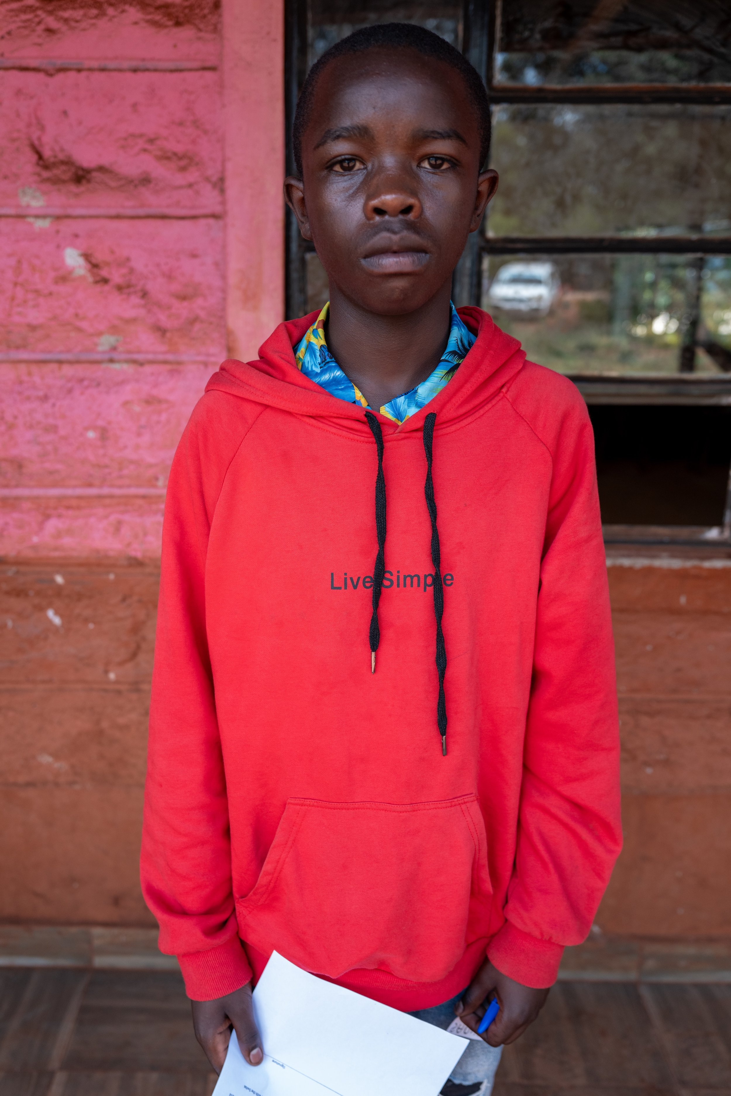 A young boy with dark skin and short hair stands indoors in front of a weathered pink wooden wall. He is wearing a red hoodie with the words 'Live Simple' printed on the chest and a blue floral shirt underneath. He holds a piece of paper and a pen in