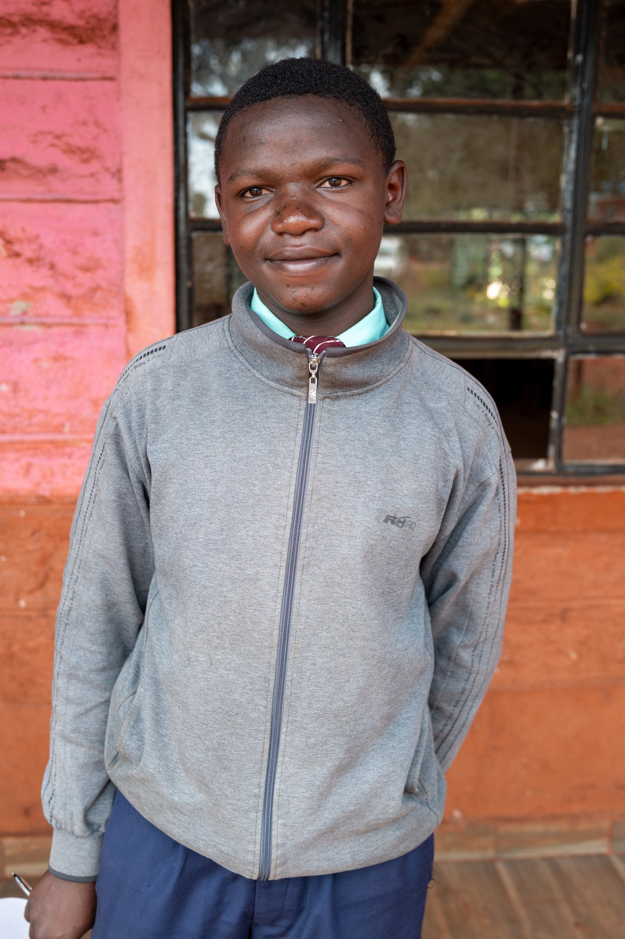 A young boy in a gray jacket and school uniform standing outdoors in front of a pink painted wall and window.