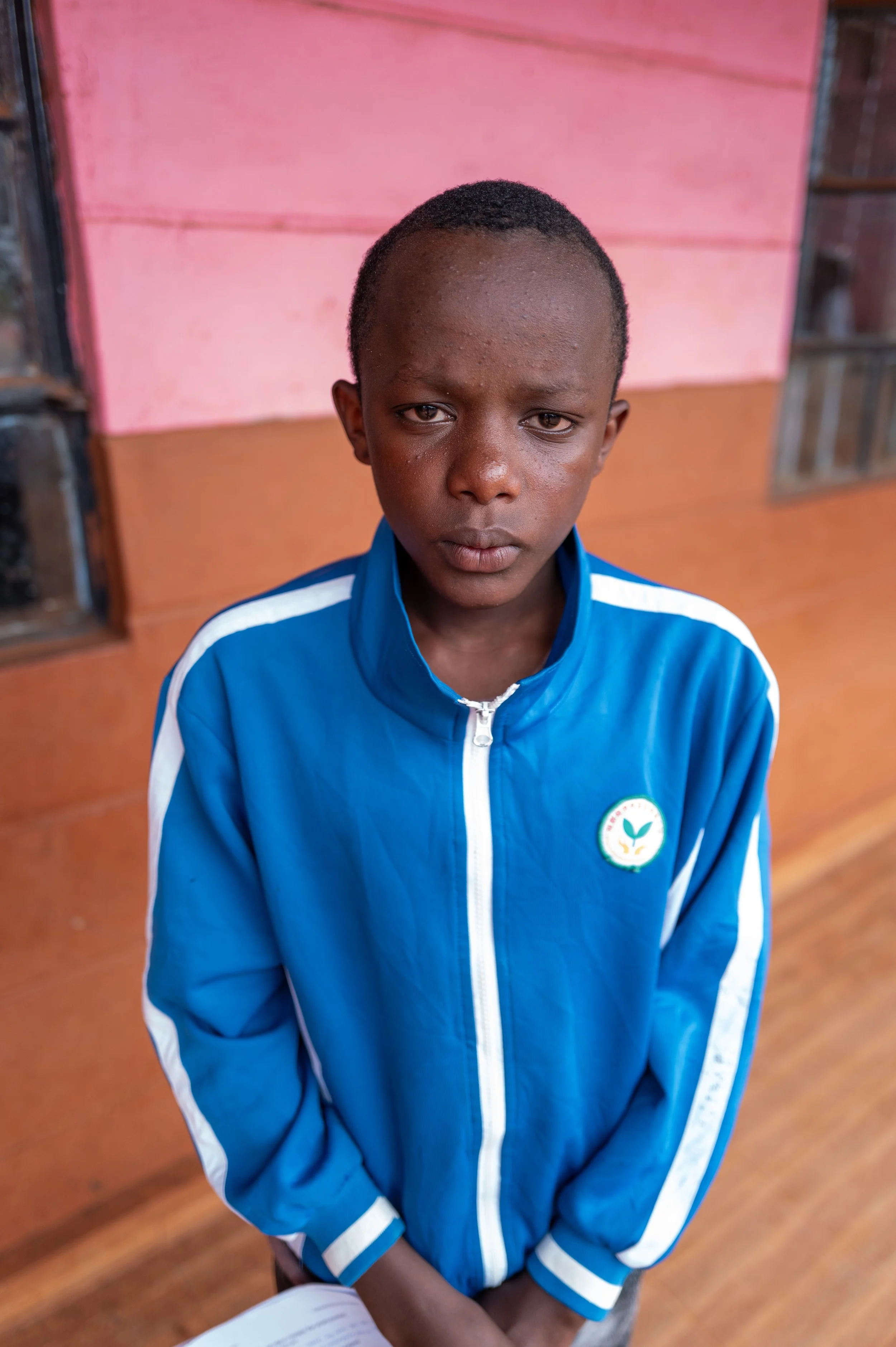 A young boy with a serious expression, wearing a blue jacket with a school or organization emblem on it, standing indoors against a pink and orange wall.