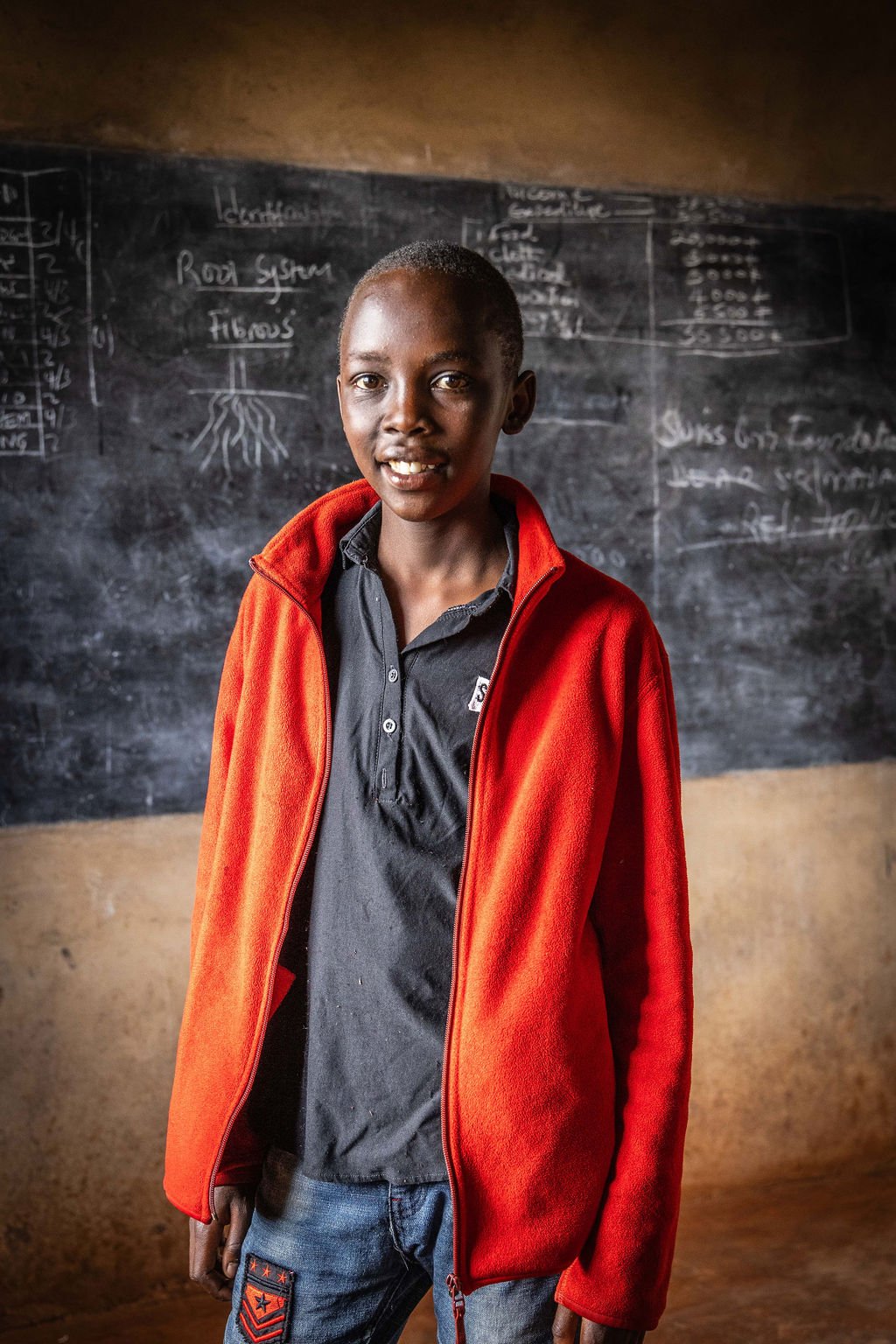 A young person standing indoors in front of a chalkboard, wearing a red jacket and black shirt. The chalkboard has diagrams and notes written on it.