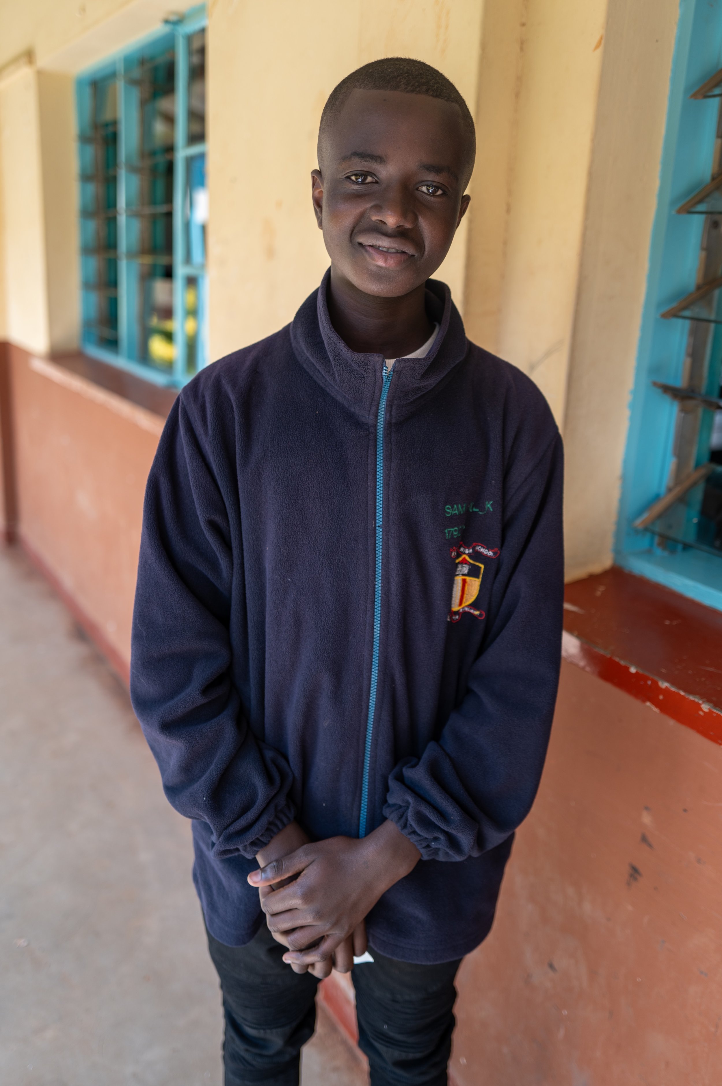 A young boy standing outdoors near a school building with yellow walls and blue windows, wearing a navy blue jacket with a crest.