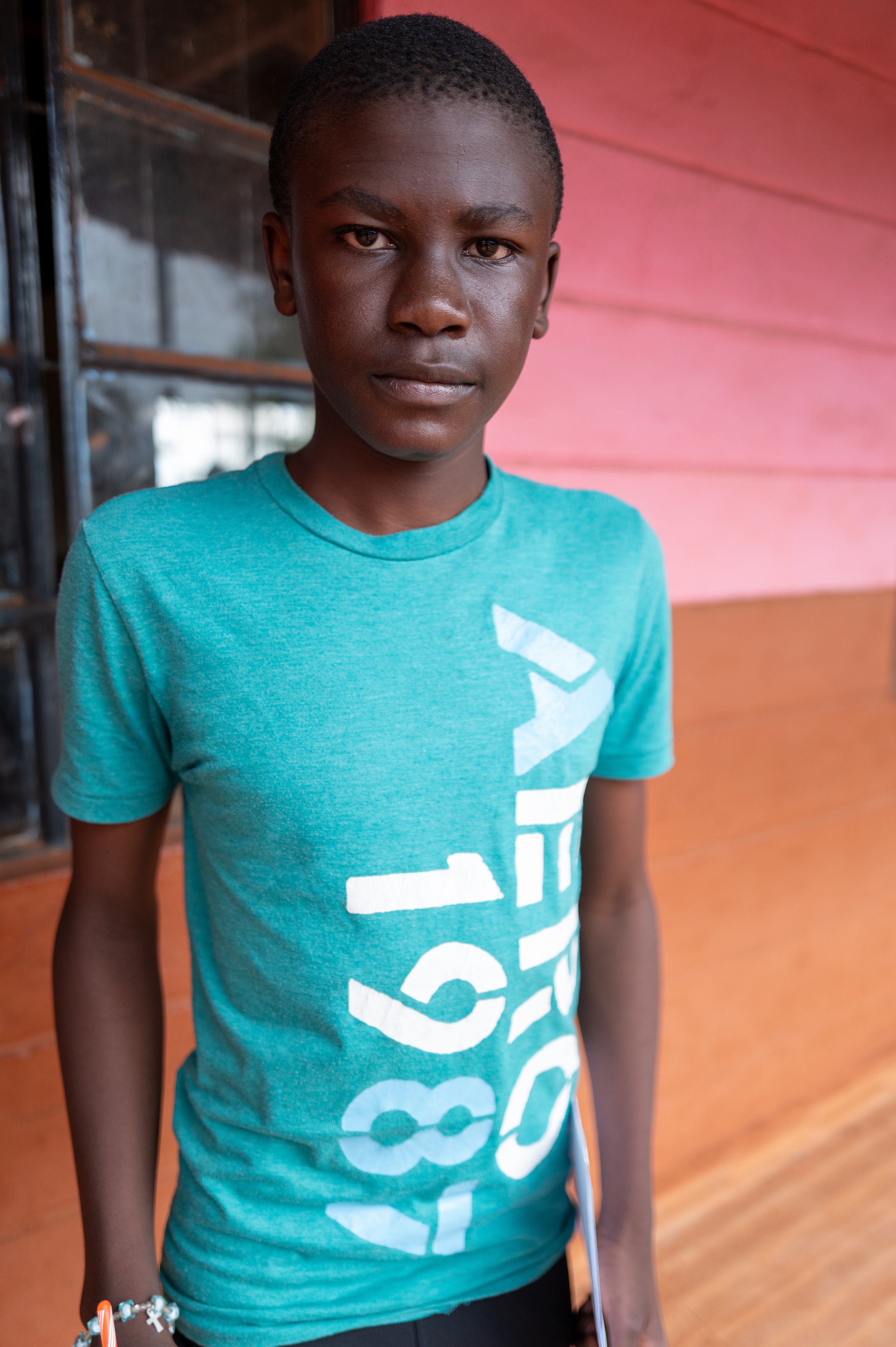 A young boy with short hair and dark skin, wearing a turquoise T-shirt with white text, standing indoors near a window and pink wall.