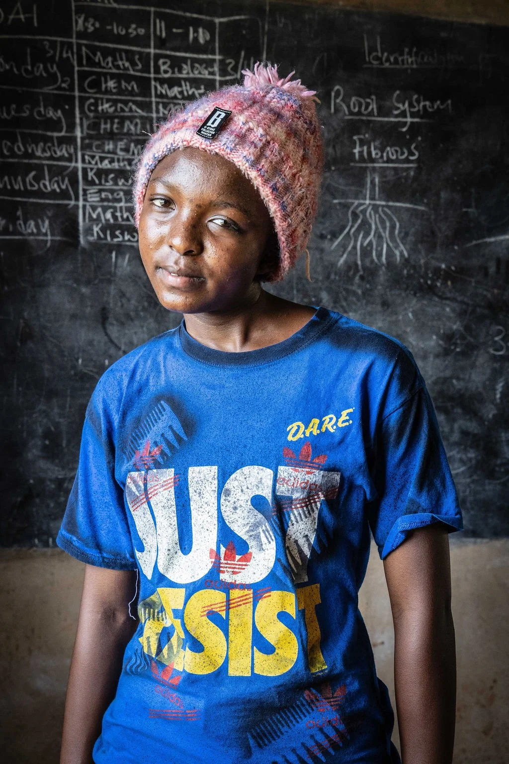 Person in a blue "Just Resist" T-shirt and pink knitted hat standing in front of a chalkboard with writing and drawings.