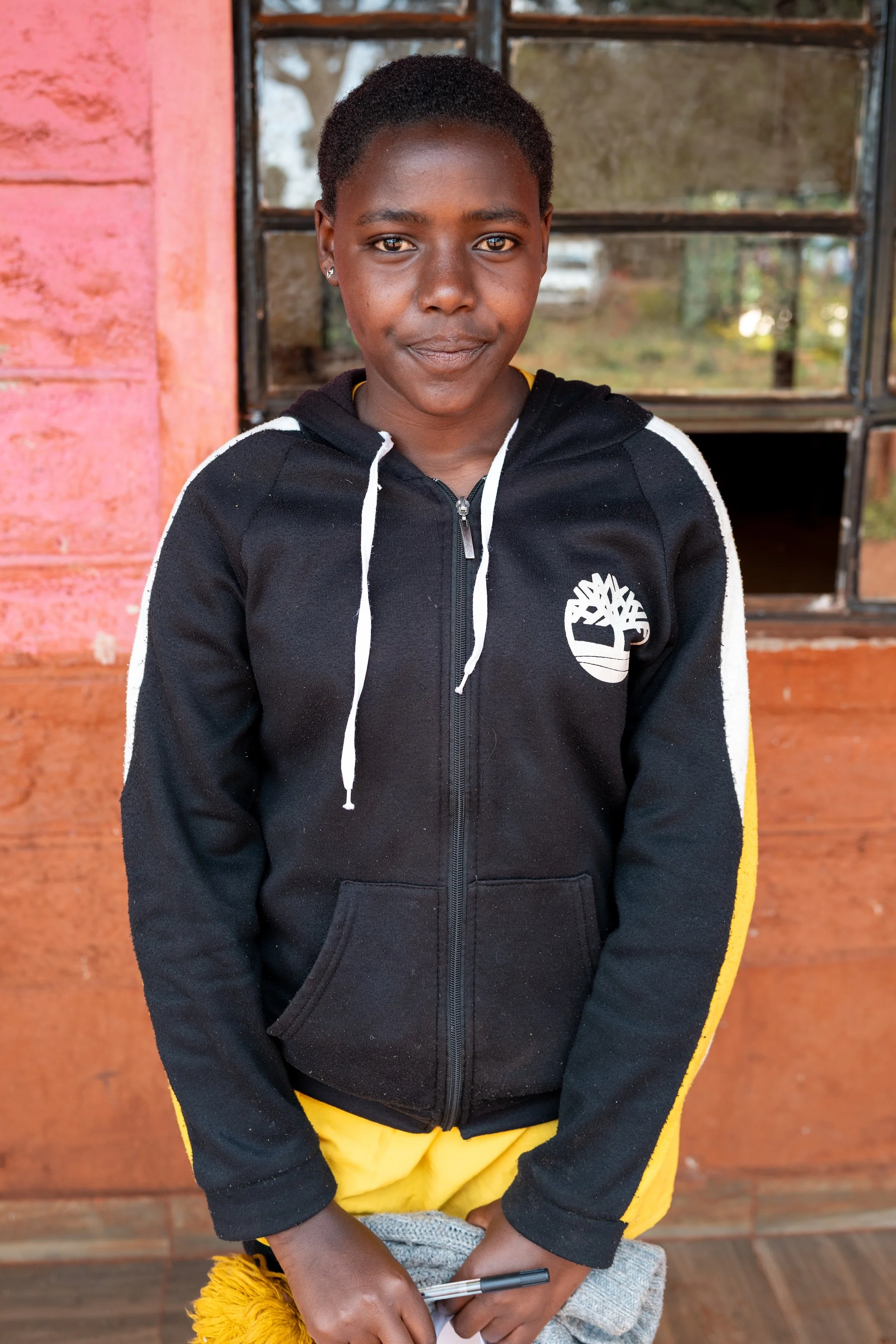 Young girl standing indoors in front of a window, wearing a black zip-up hoodie with white and yellow accents, holding a gray cloth and pen, looking at the camera.