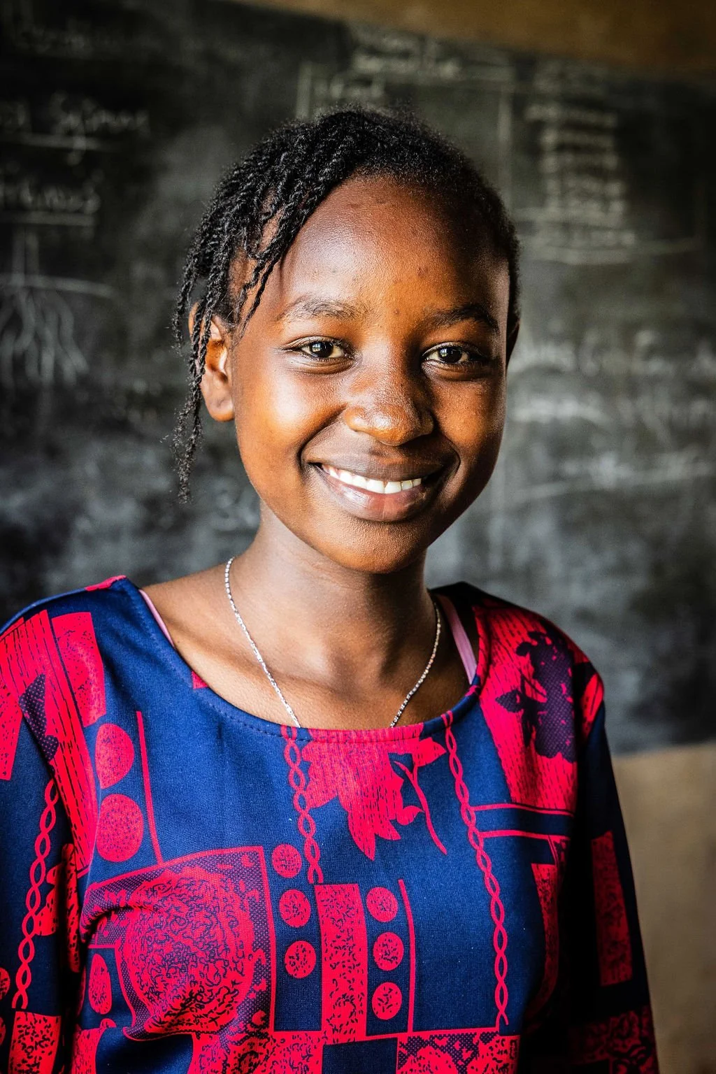 Smiling girl in colorful dress in front of a chalkboard.