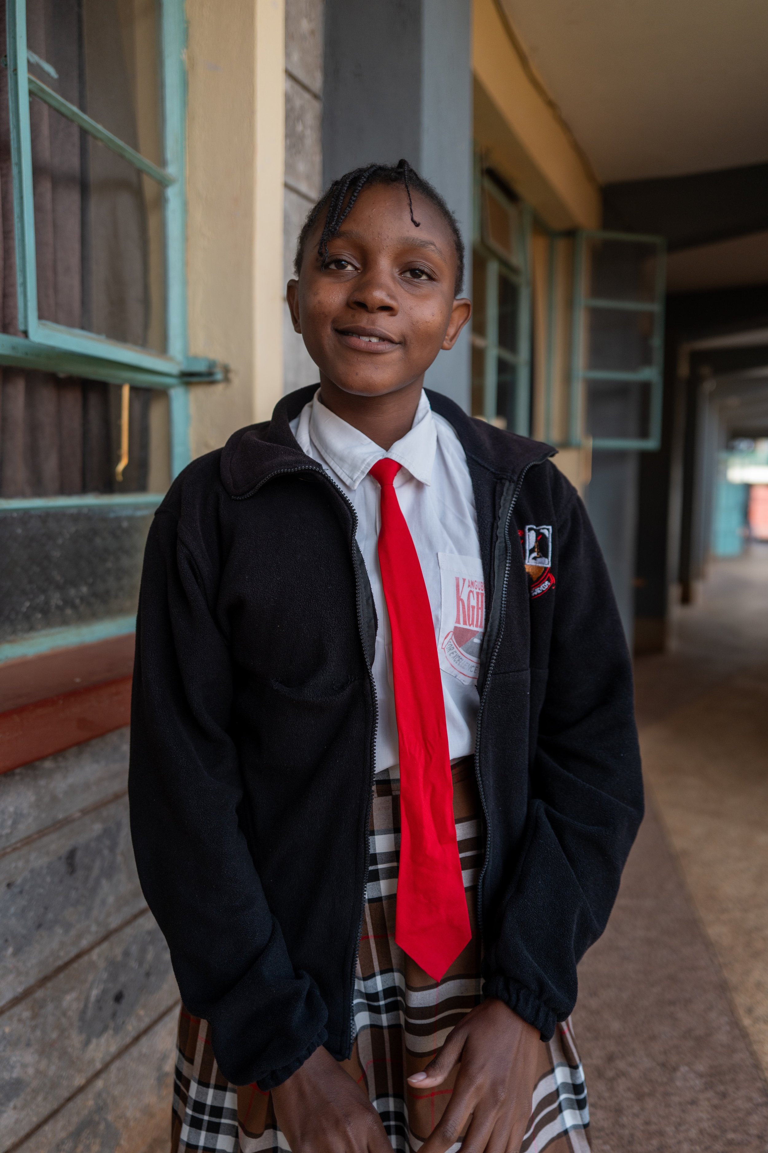 A young girl in school uniform, wearing a white shirt with a red tie, a plaid skirt, and a black jacket, stands outdoors near a building with open windows and smiling at the camera.