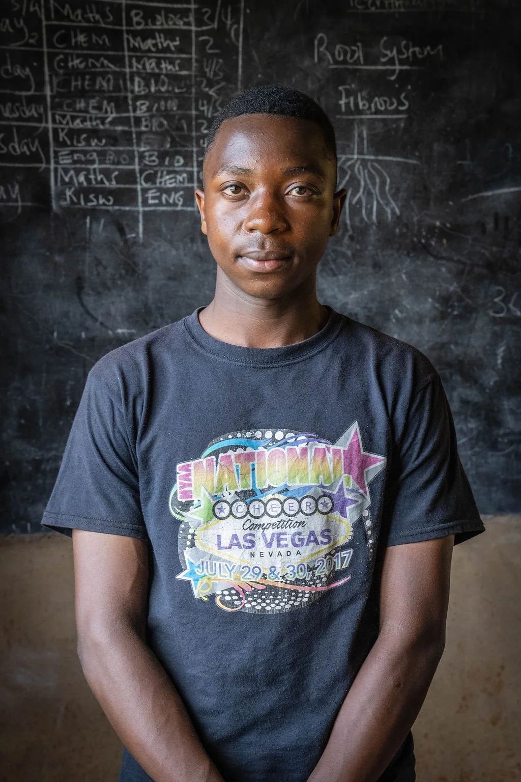 A person wearing a black t-shirt with colorful text about a cheer competition in Las Vegas stands in front of a blackboard with a schedule and notes.