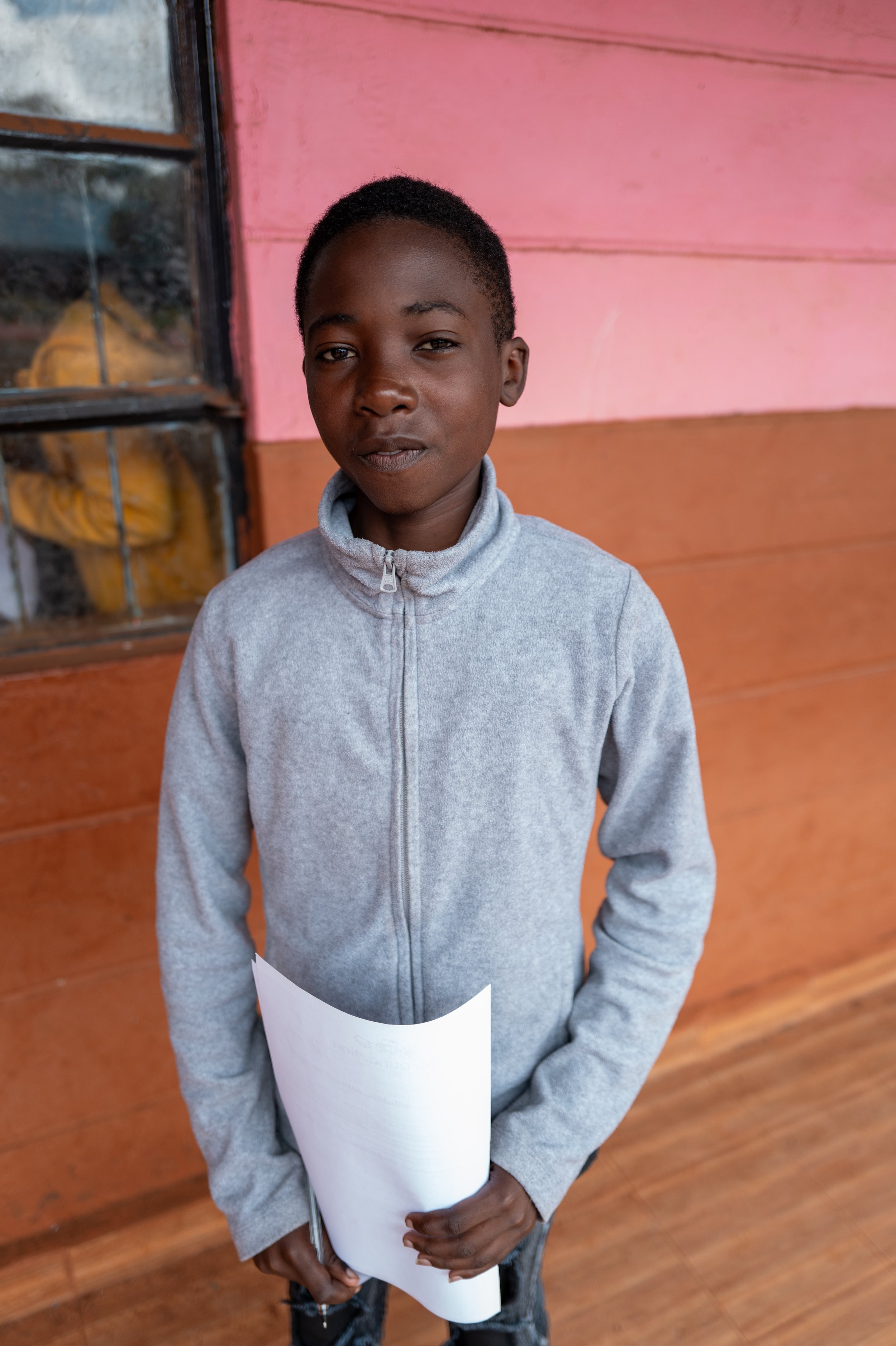 A young boy in a gray zip-up jacket holding a white piece of paper or notebook, standing in front of a colorful wooden wall with pink, orange, and brown hues.