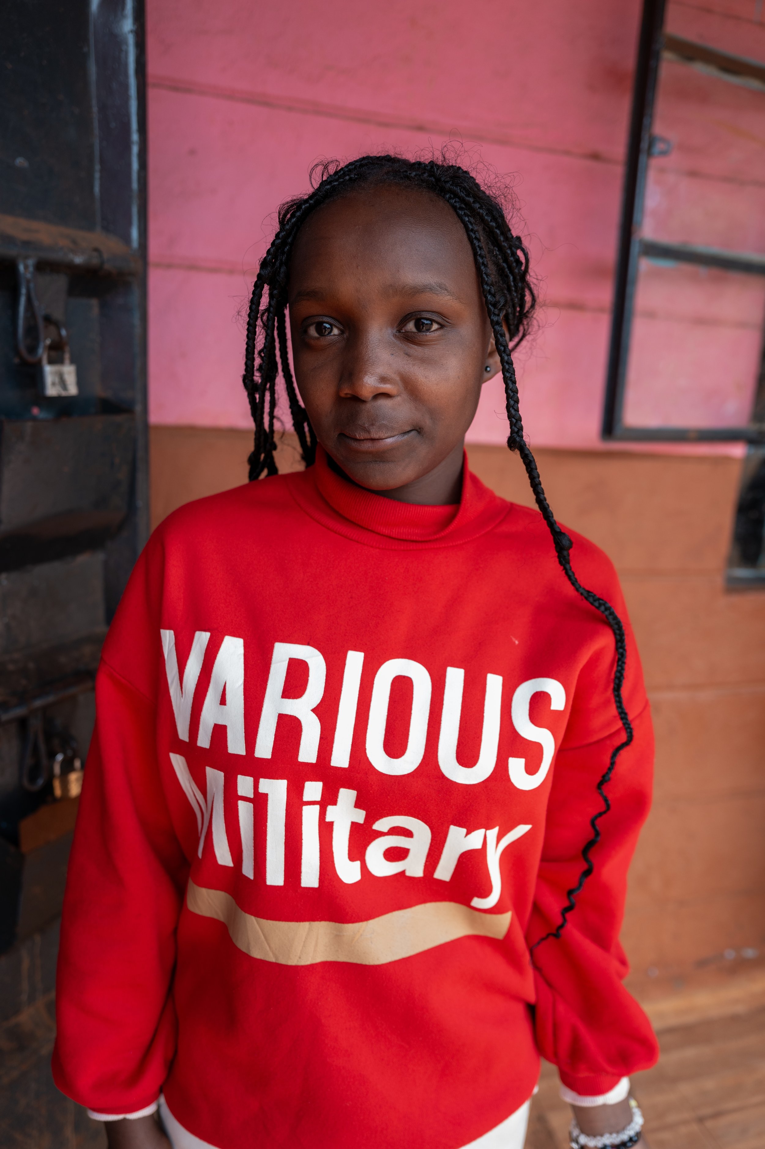 A young girl with braided hair wearing a red sweatshirt that says 'VARIOUS MILITARY' standing indoors against pink and brown walls.