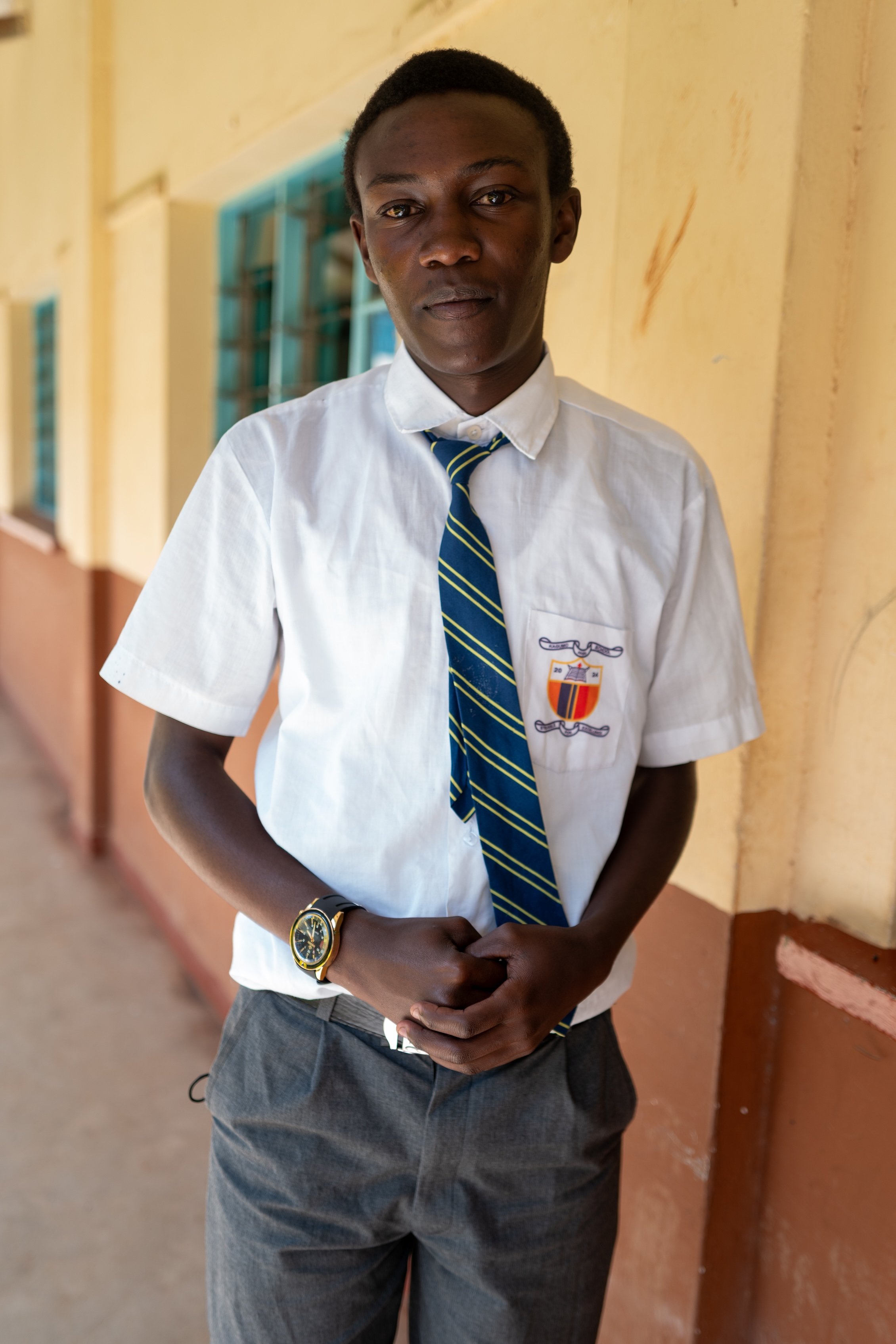 A young man in a school uniform standing in a corridor, wearing a white shirt, navy blue and yellow striped tie, gray pants, and a wristwatch.