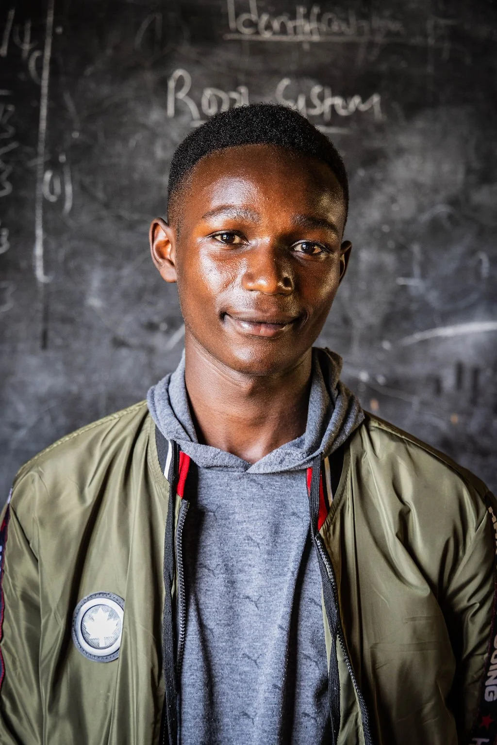 Young man wearing a green jacket and gray hoodie, standing in front of a blackboard.