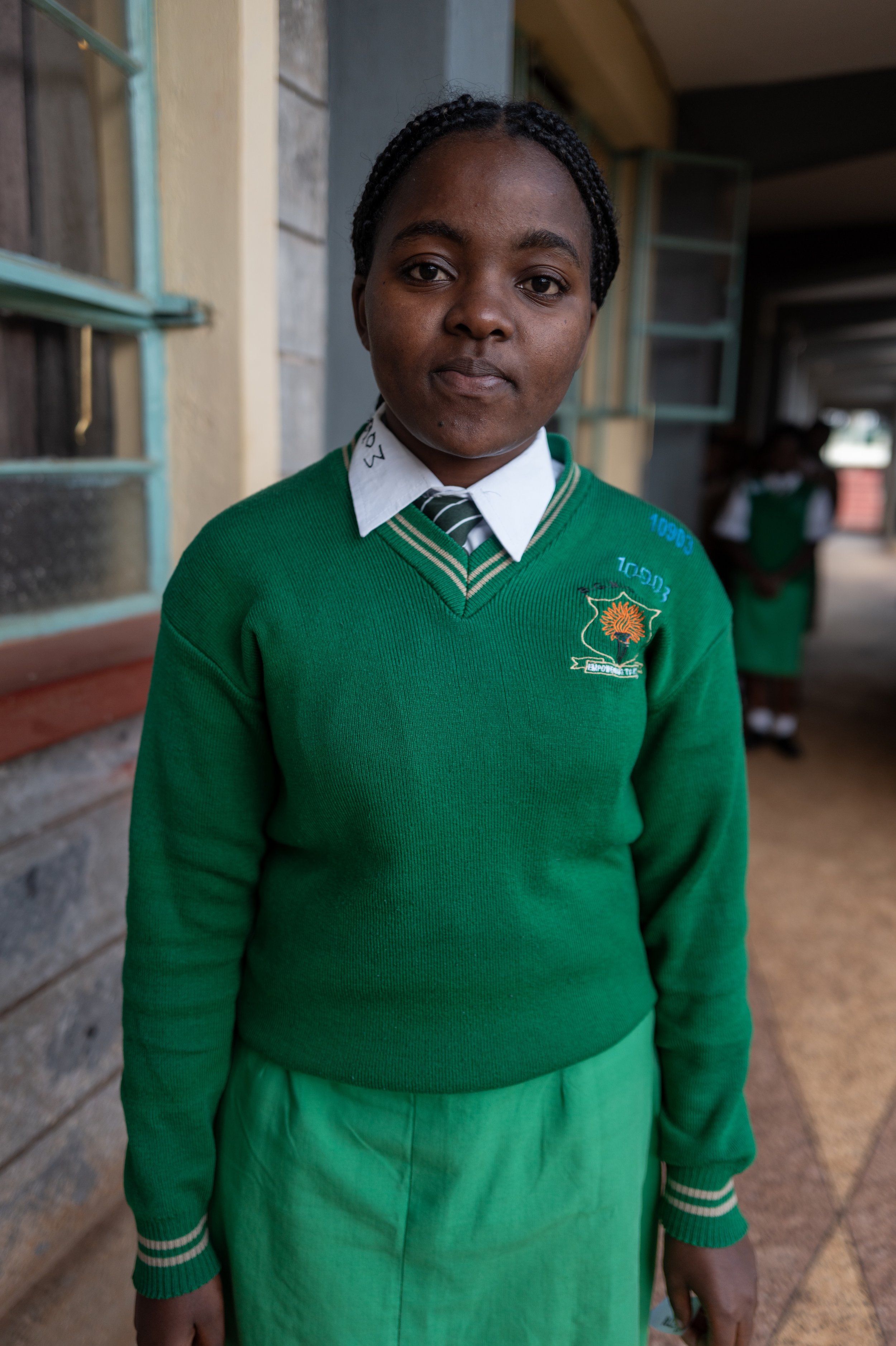 A young girl wearing a green school uniform with a white shirt, striped tie, and a green sweater with an embroidered emblem. She stands outside near a building with a window and other students in the background.