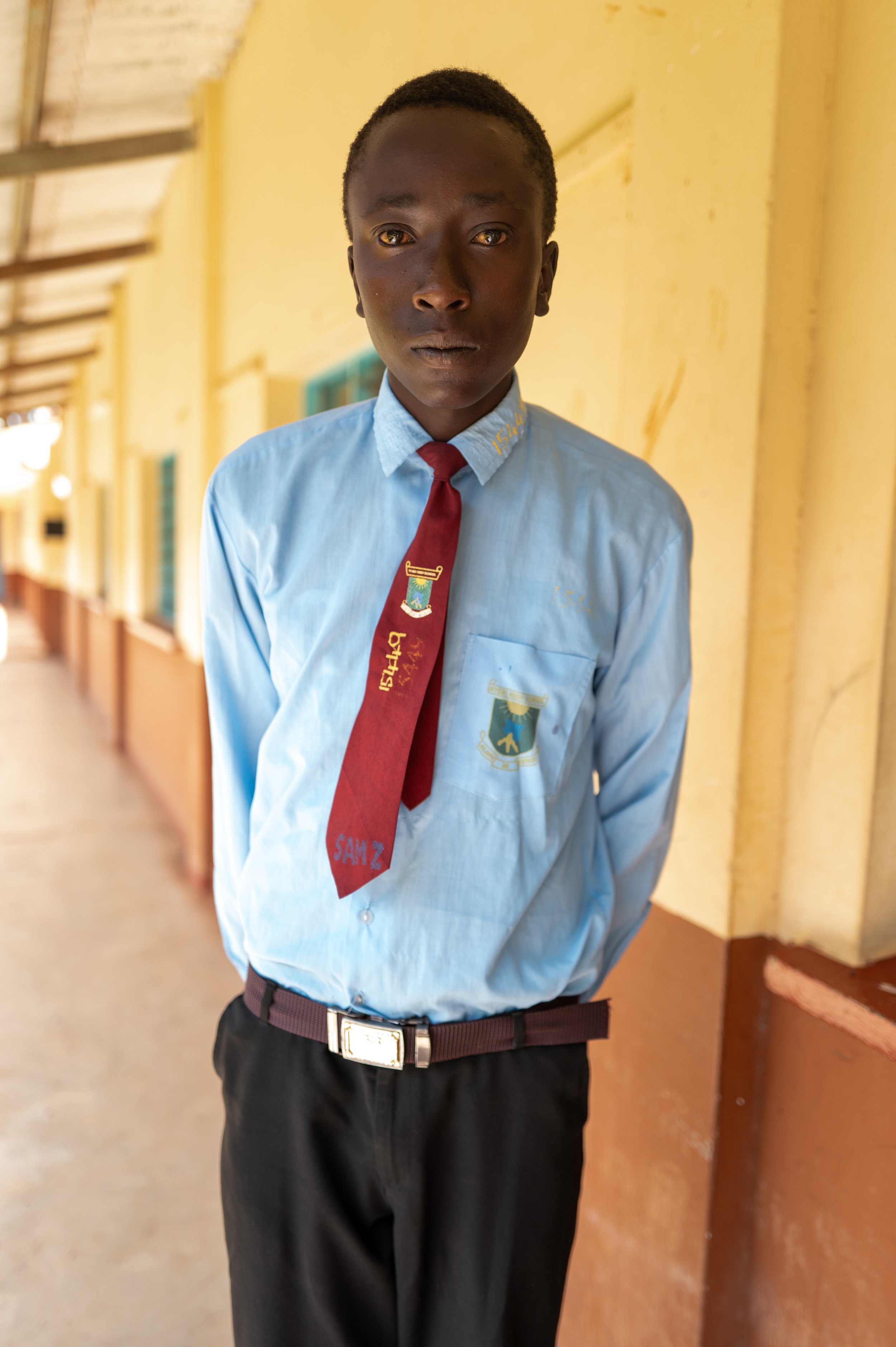 A schoolboy in a light blue shirt with school emblem, red tie, and black trousers standing in a school corridor.