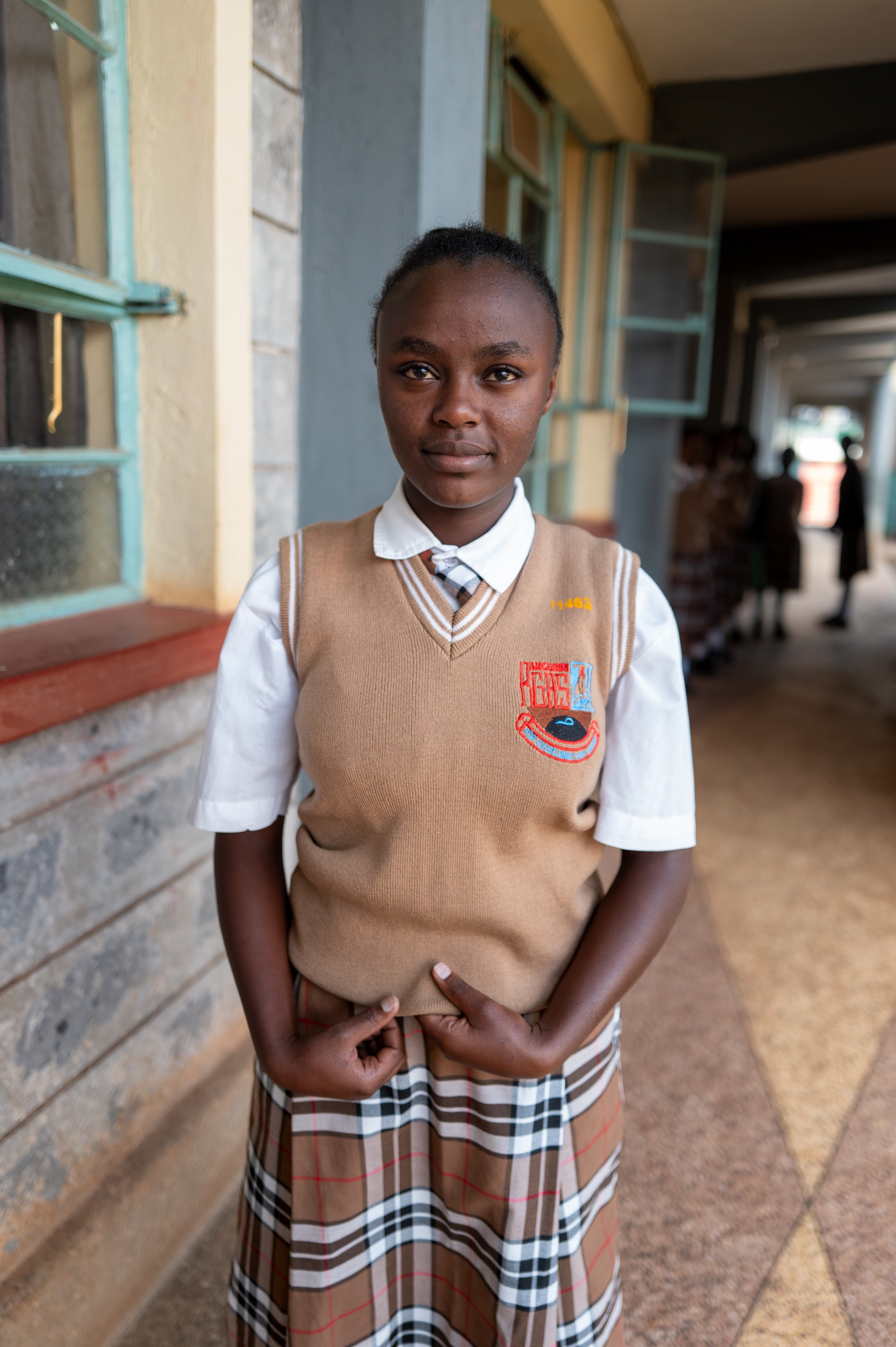 A young girl wearing a school uniform standing outside a building with open windows, looking at the camera.