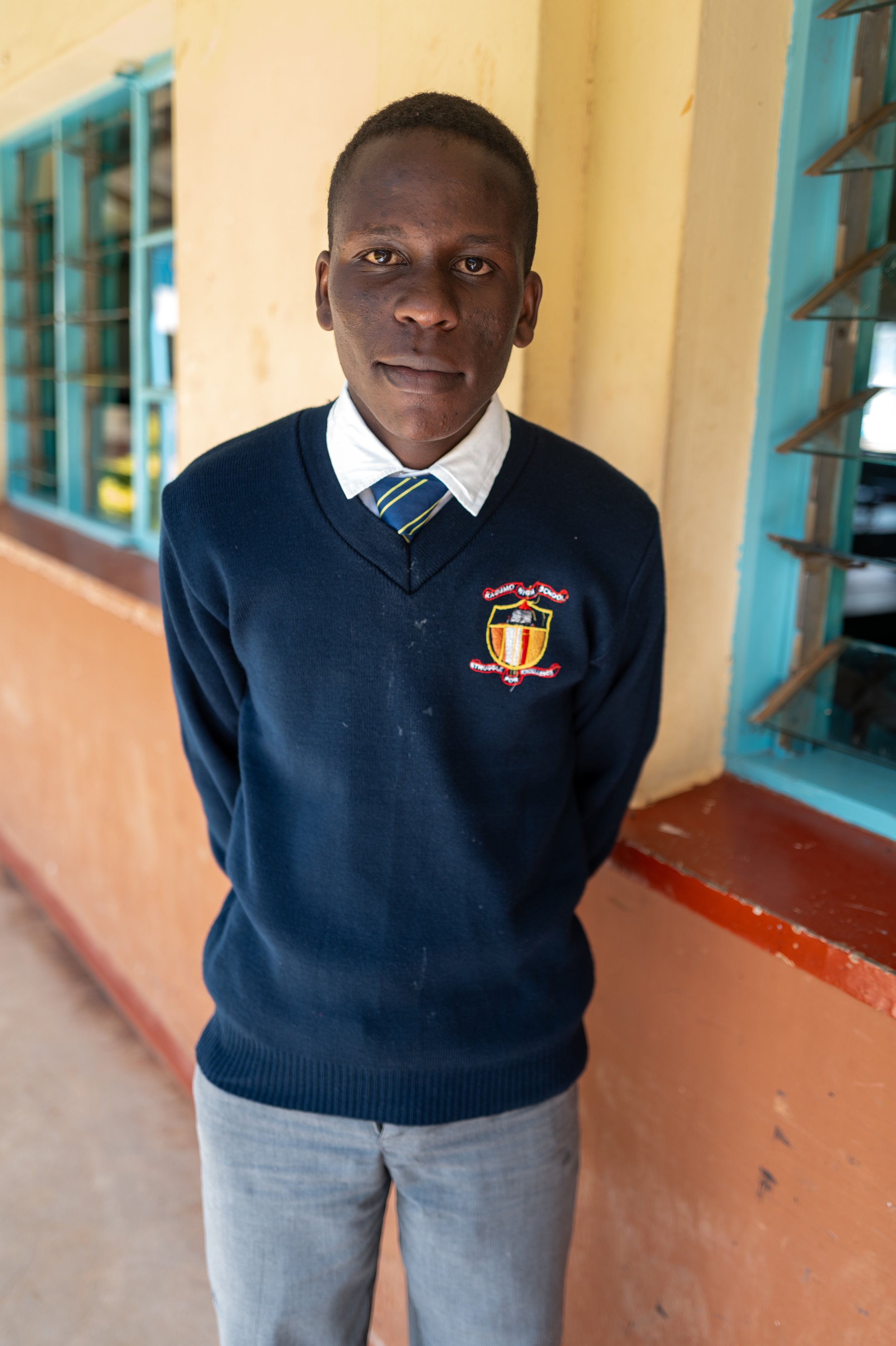 A young male student in a school uniform standing in front of a yellow wall and window with blue frames.