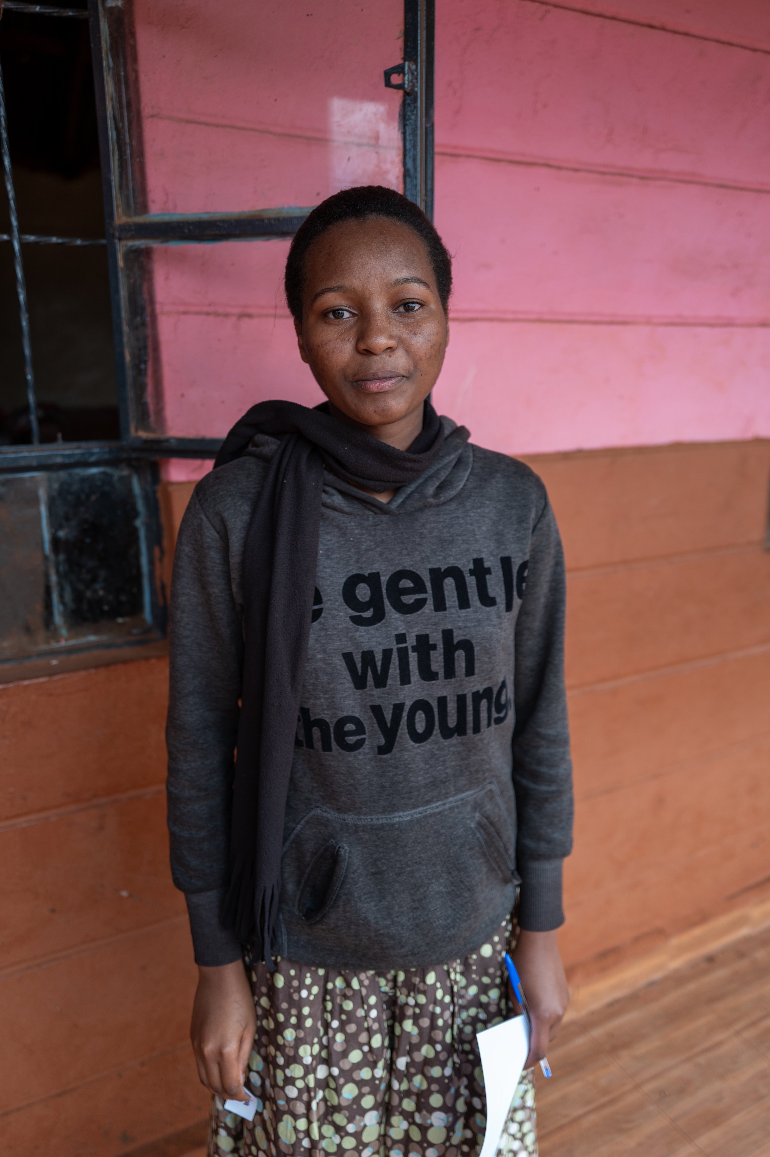 A young girl with short hair, wearing a dark gray hoodie with the words 'gentle with the young' printed in black, standing indoors in front of a pink wall with black window frames. She is holding a small notebook or paper and a pen in her right hand.
