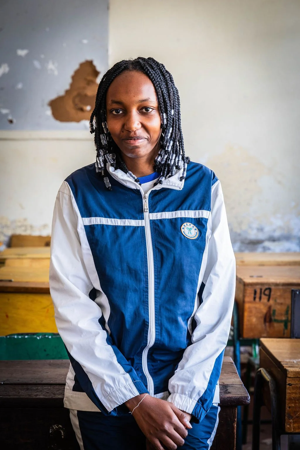 A person with braided hair and beaded ends is standing in a classroom, wearing a blue and white jacket.