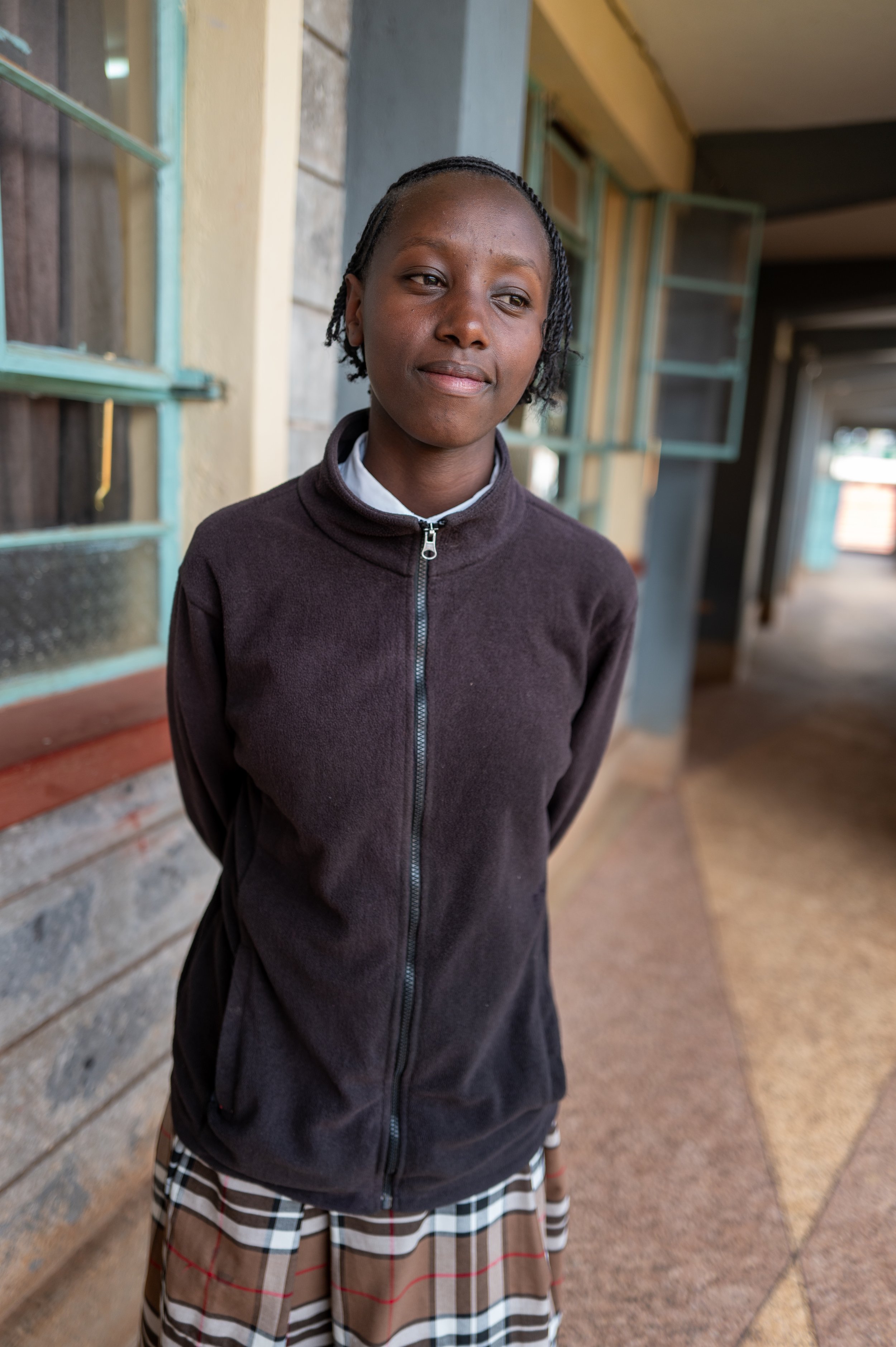 A young girl with braids standing outside a building with open windows, looking to the side with a neutral expression, wearing a dark zip-up jacket and a plaid skirt.