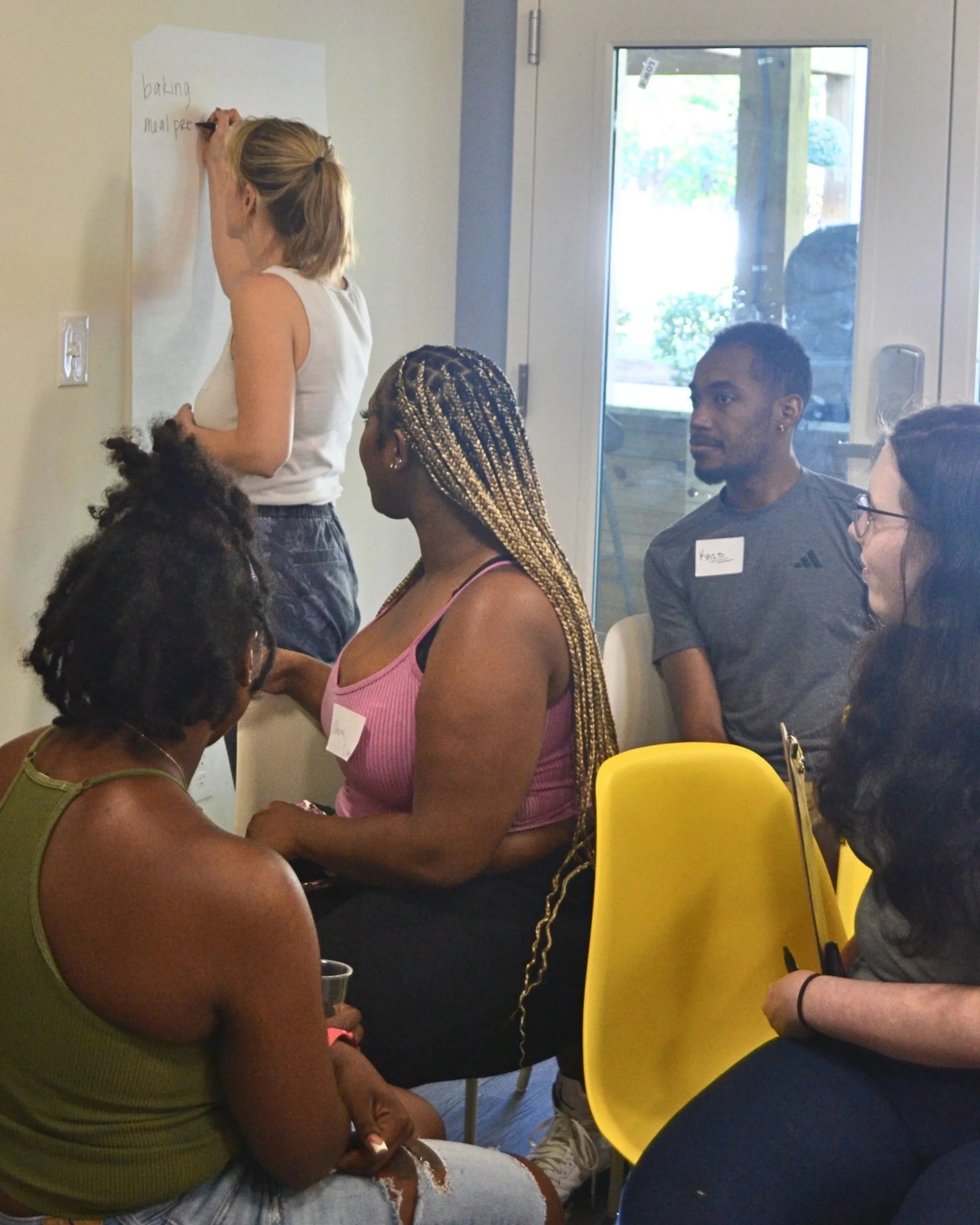 Kait of Burkett Farm writing brainstormed food topics on a wall with Kitchen Compass participants looking on.