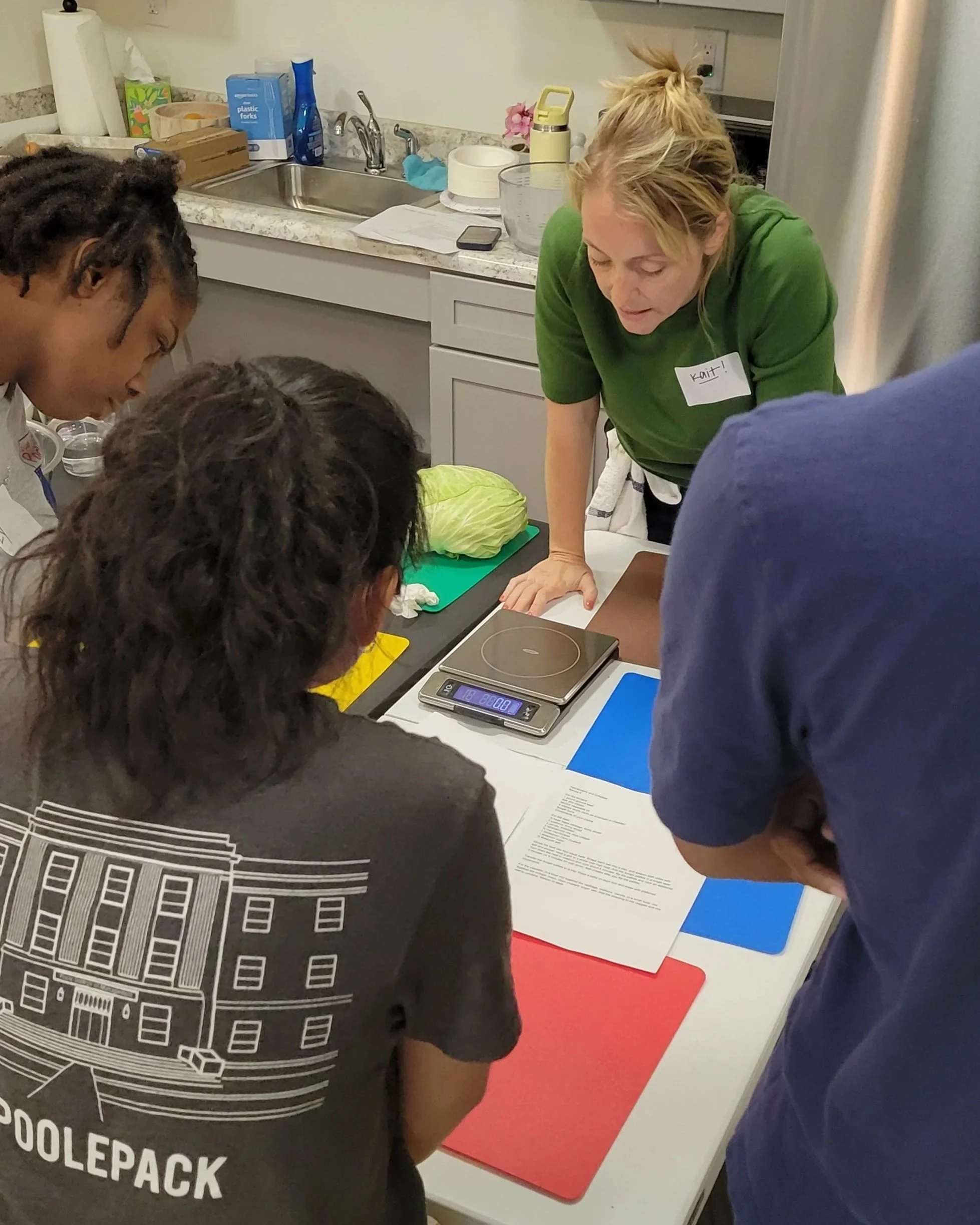 Three Kitchen Compass clients and Kait from Burkett Farm leaning over a table with cutting boards to look at recipes together. 