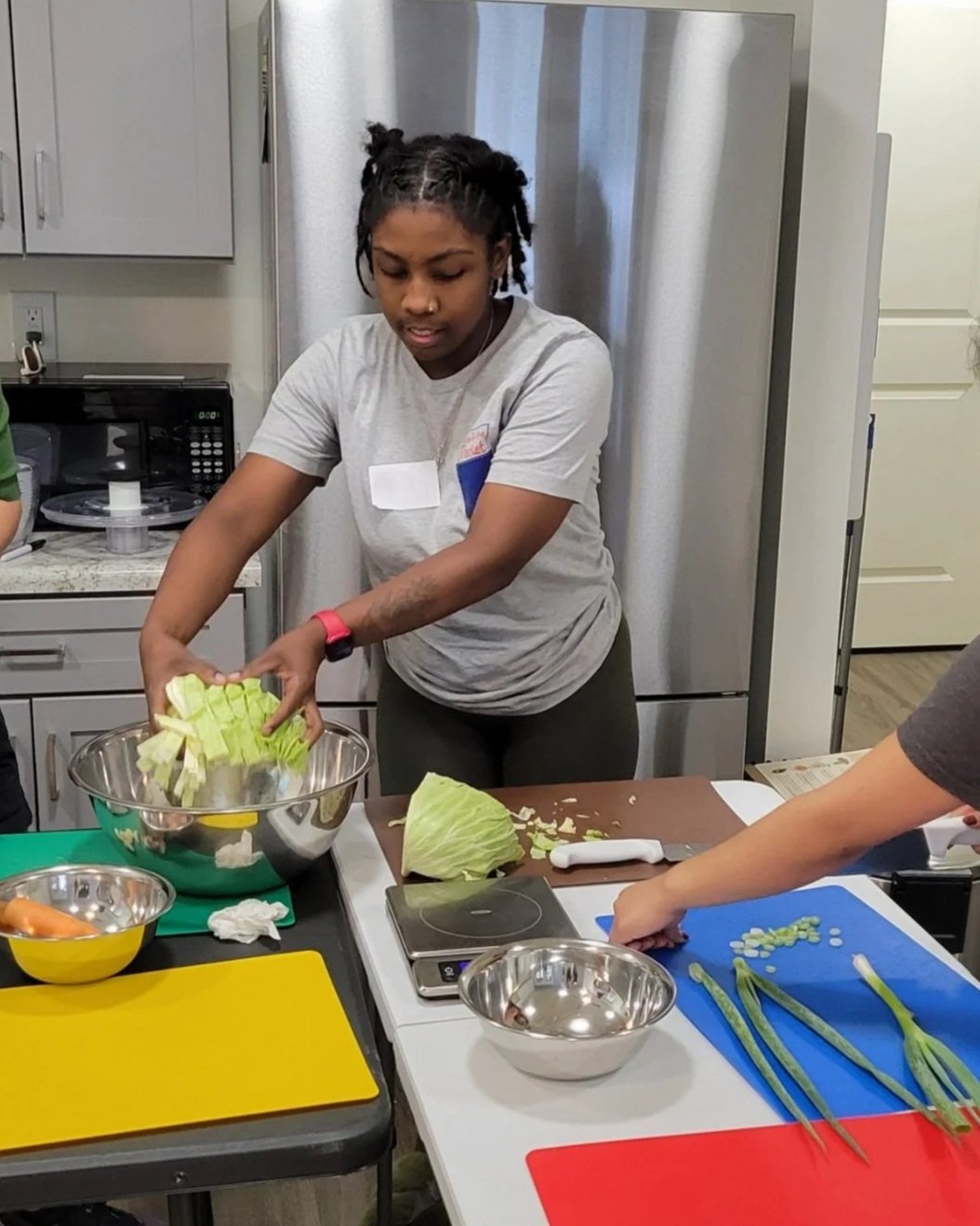 A Kitchen Compass client dropping sliced cabbage into a mixing bowl. Another client is chopping green onions.