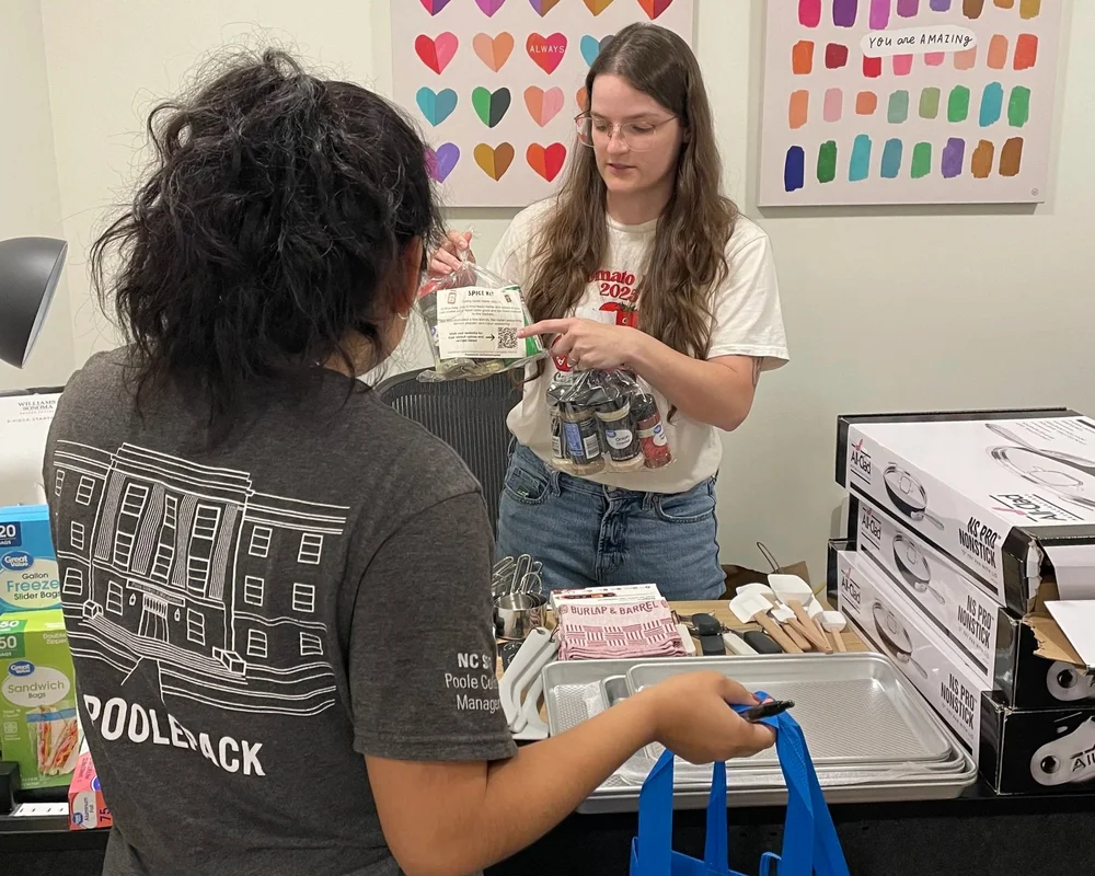 Tori of Food Equity Collaborative holding two spice kits showing a Kitchen Compass client what's inside. On the table are many kitchen tools for clients to take home, such as pans, kitchen towels, spatulas, and measuring cups.