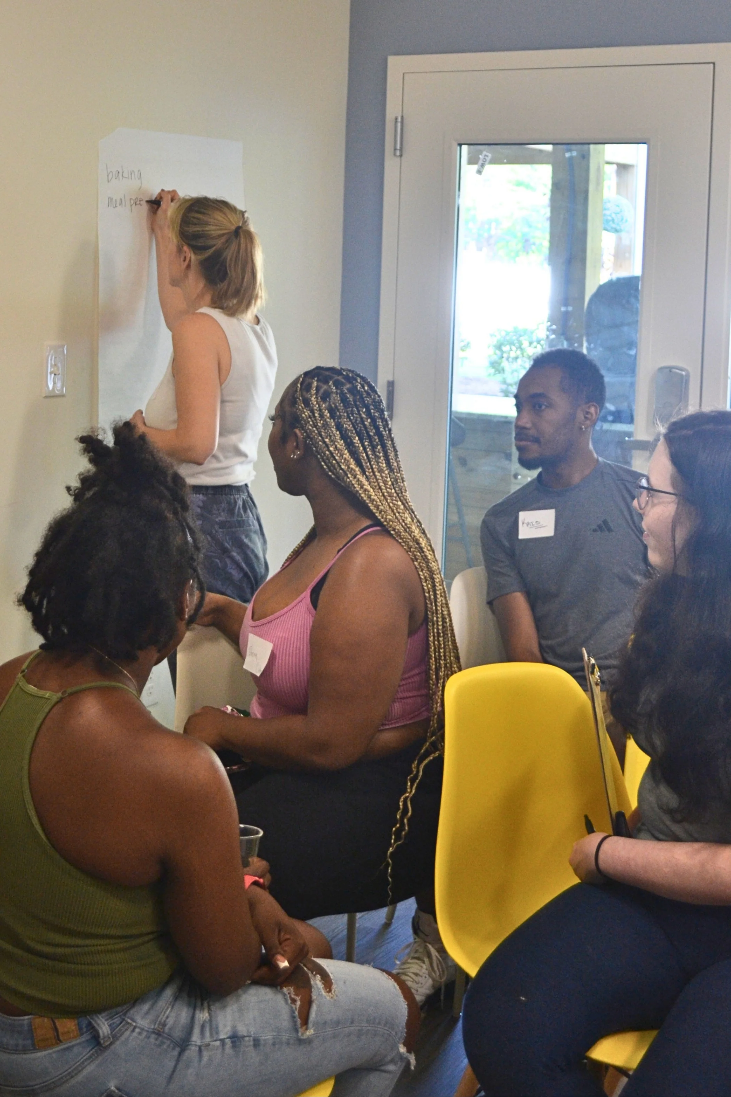 Kait of Burkett Farm writing brainstormed food topics on a wall with Kitchen Compass participants looking on.