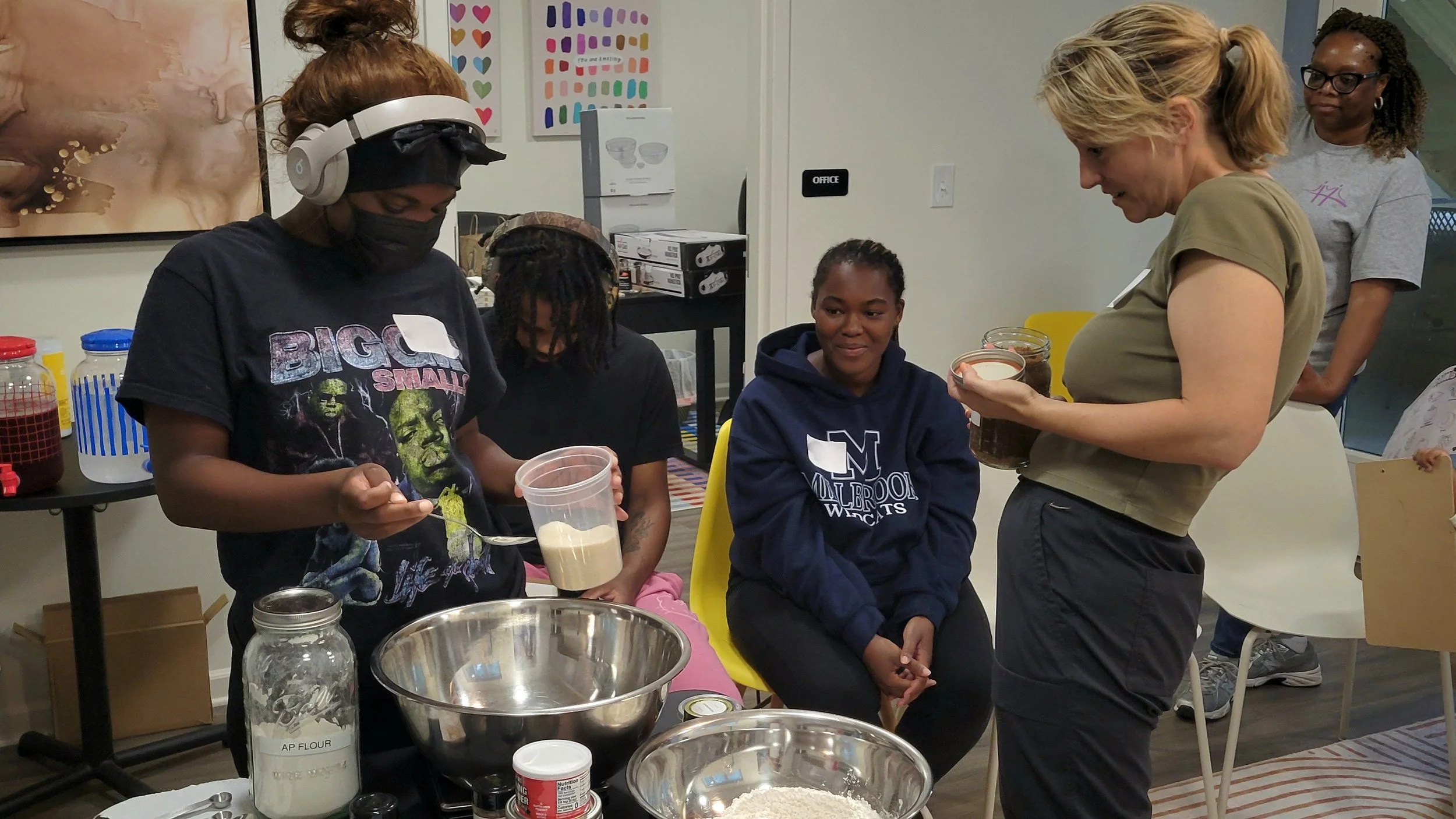 A woman measuring dry ingredients into a mixing bowl using a scale while others look on.