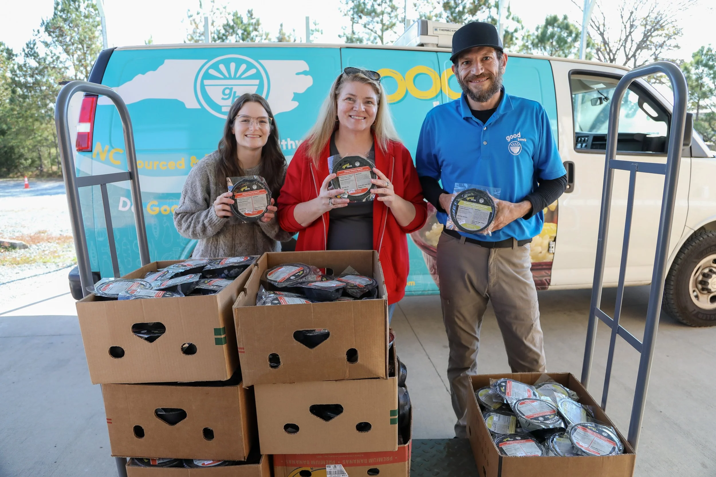 Two women, Tori Wierzchowski of Food Equity Collaborative and Melissa Driver Beard of CORA Food Pantry, and one man, Steve Navarra of Equiti Foods, stand miling in front of the Good Bowls van and behind a cart, each holding a Good Bowl. On the cart are 7 cardboard boxes full of Good Bowls.