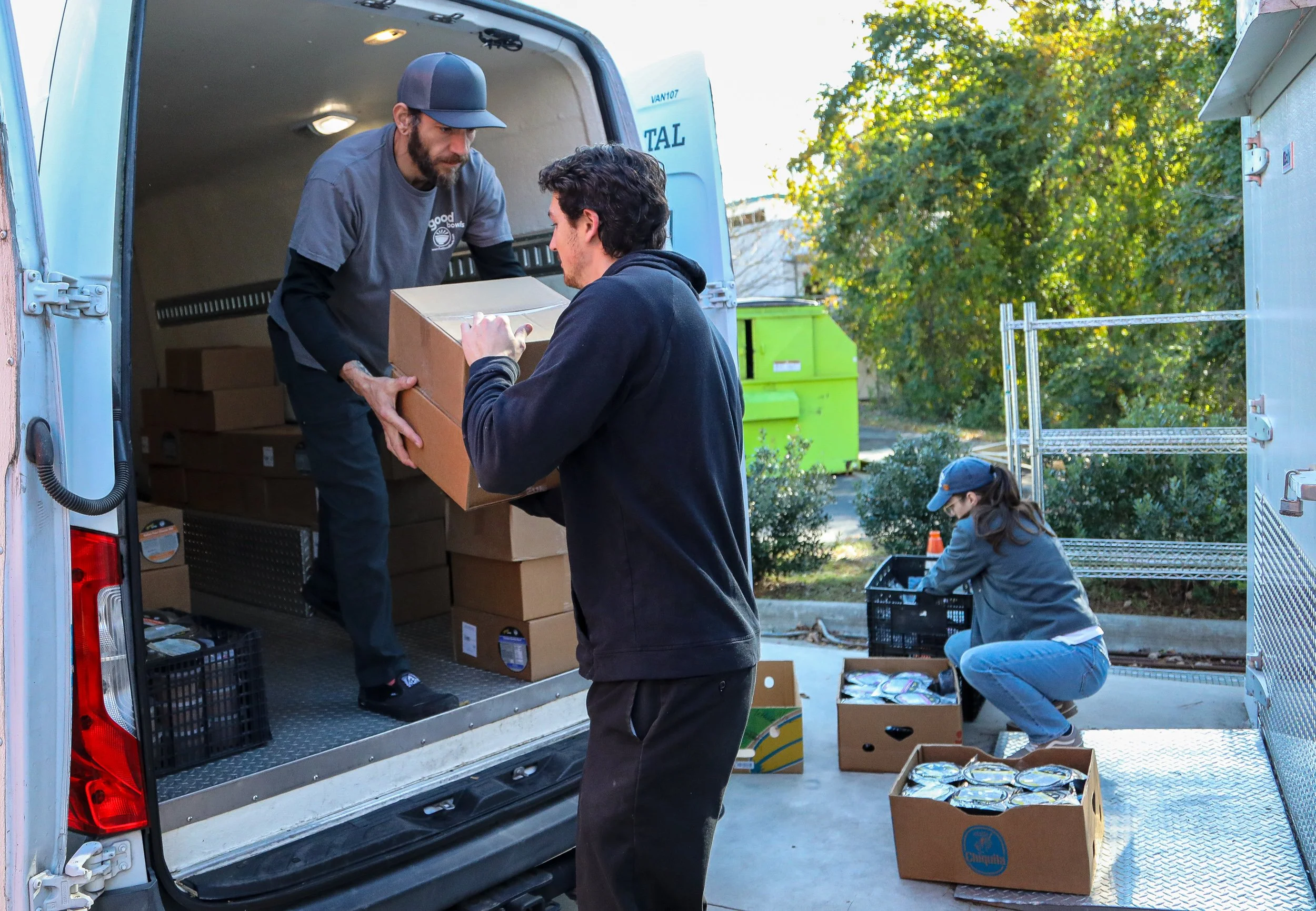 Two men stand at the back of the van full of boxes of Good Bowls. One man, Steve Navarra of Equiti Foods, is handing two boxes to another man, a volunteer at CORA Pittsboro. In the background, a woman, Tori Wierzchowski, Executive Director of Food Equity Collaborative, is crouched down on the ground moving Good Bowls from plastic bins to cardboard boxes.