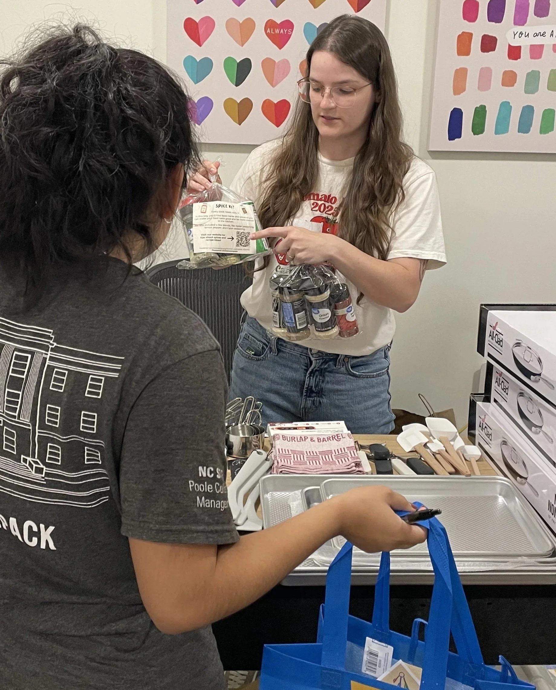 FEC Executive Director showing a client their new spice kit standing over a table with kitchen tools to pick from, including baking sheets, pans, kitchen towels, whisks, spatulas, and more.